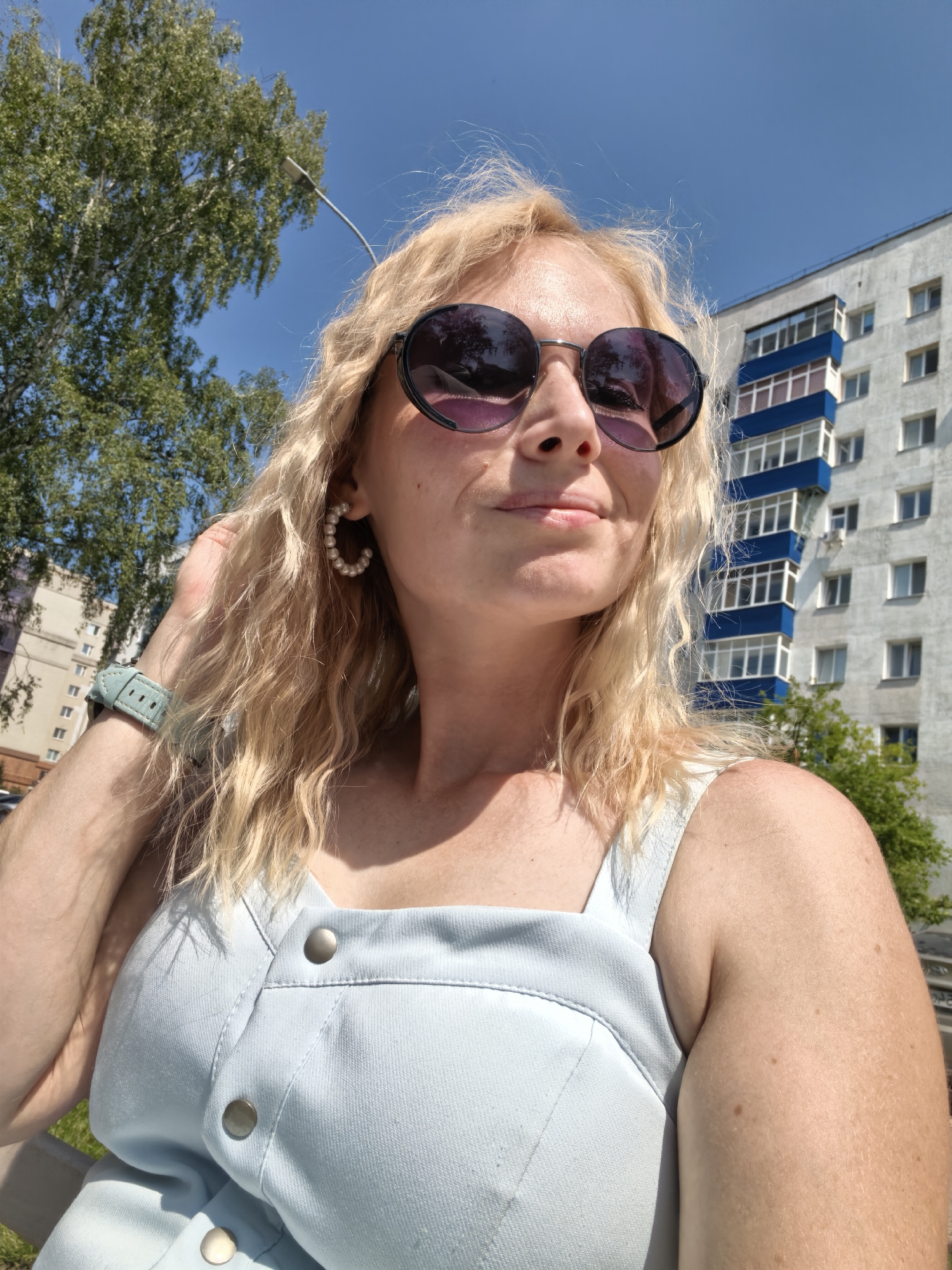 A blonde woman wearing sunglasses, pearl hoop earrings, and a light blue dress is taking a selfie outdoors. She is standing in front of a multi-story residential building with blue and white accents. A large tree with green foliage is visible to the left of the frame, and the sky is clear blue. The woman's left arm is resting on her lap, and she is wearing a light blue watch. The scene appears to be from Sterlitamak, Russia.