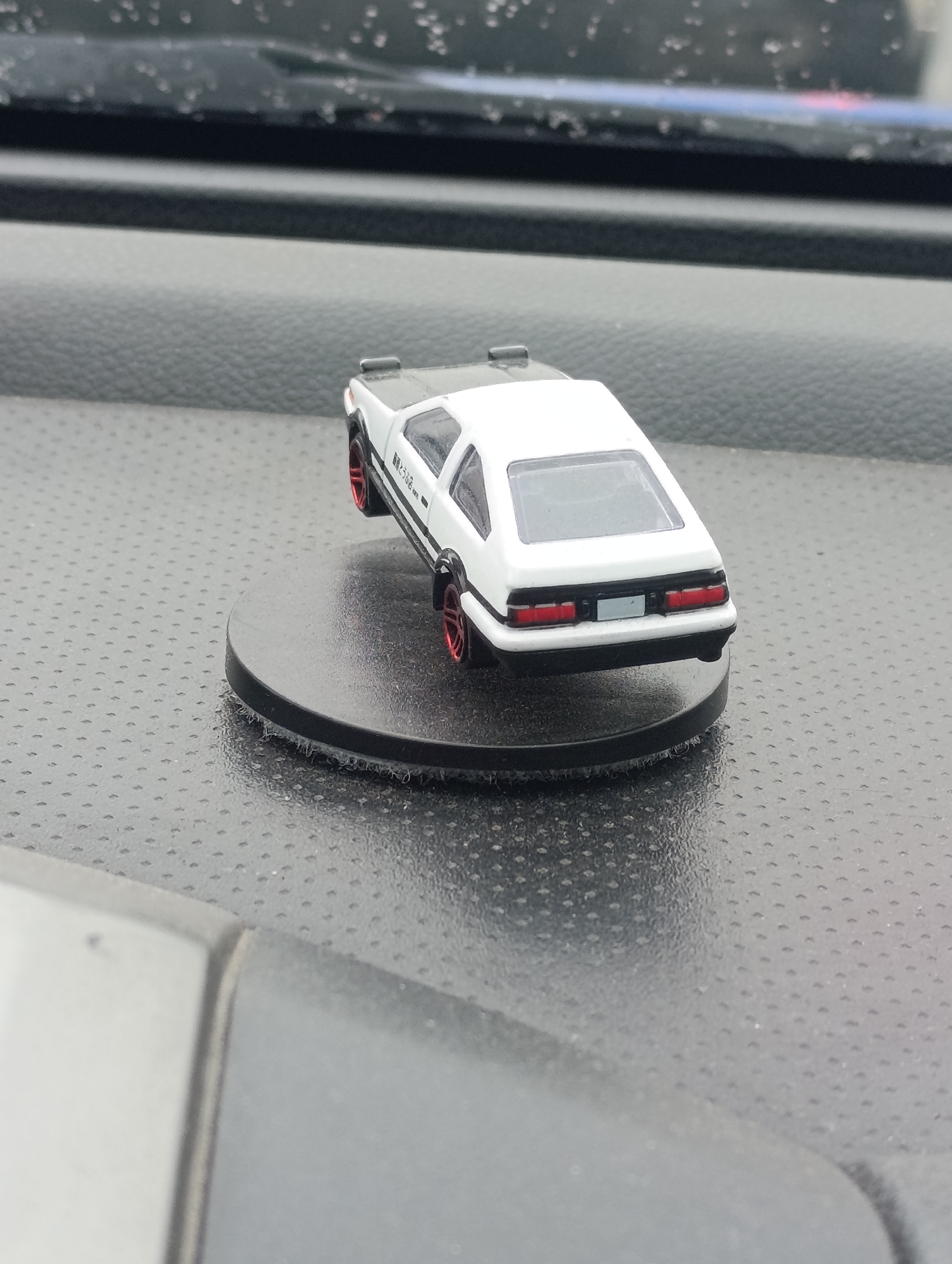 The image displays a miniature white Toyota AE86 Sprinter Trueno model car, characterized by its black hood, red wheel rims, and black Japanese text "藤原とうふ店 (自家用)" (Fujiwara Tofu Shop (Private Use)) on its side. The model car is positioned on a circular black display stand. This stand rests on the textured dashboard of a vehicle, which features a dark, subtly reflective surface with a fine dotted pattern. In the background, water droplets are visible on the windshield or another surface, indicating rainy or damp weather conditions. The lighting suggests it is daytime, possibly overcast. The scene captures a static display within a car interior. No people are visible, and no activity or event is taking place beyond the stationary presentation of the miniature. The location context provided is Krasnoyarsk, Russia.