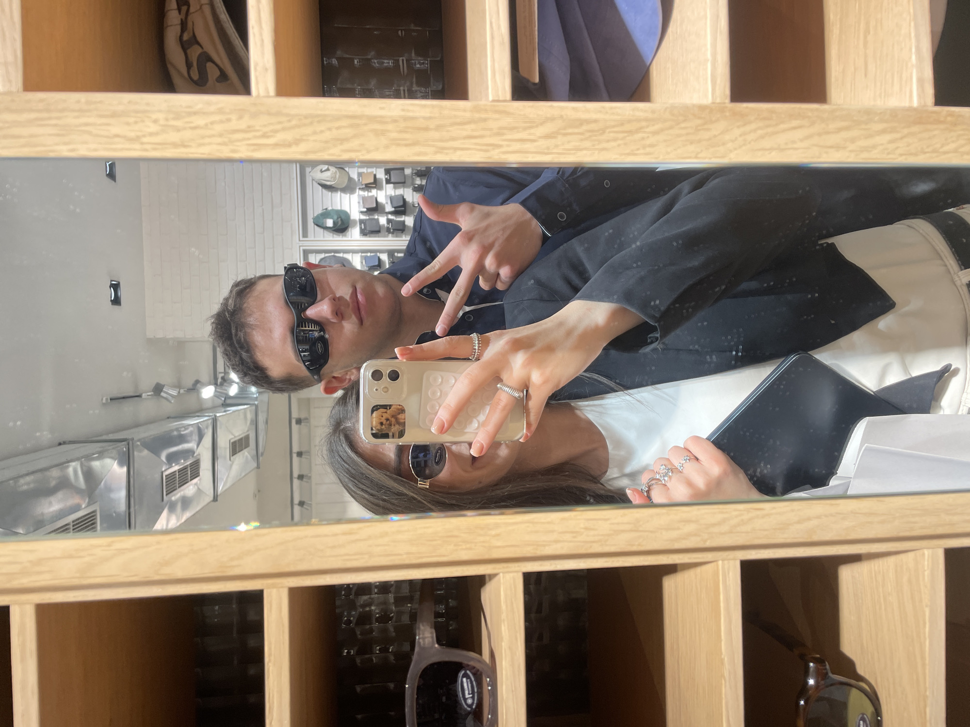 In Yerevan, Armenia, a selfie shows a man and a woman reflected in a mirror. The man wears sunglasses, a dark jacket over a collared shirt, and is making a peace sign with his fingers. The woman also wears sunglasses and holds a phone with a dog image on the case and a tablet in her other hand. They are in a store with wooden shelving displaying sunglasses and other merchandise. The background reflects store lights and ventilation units.