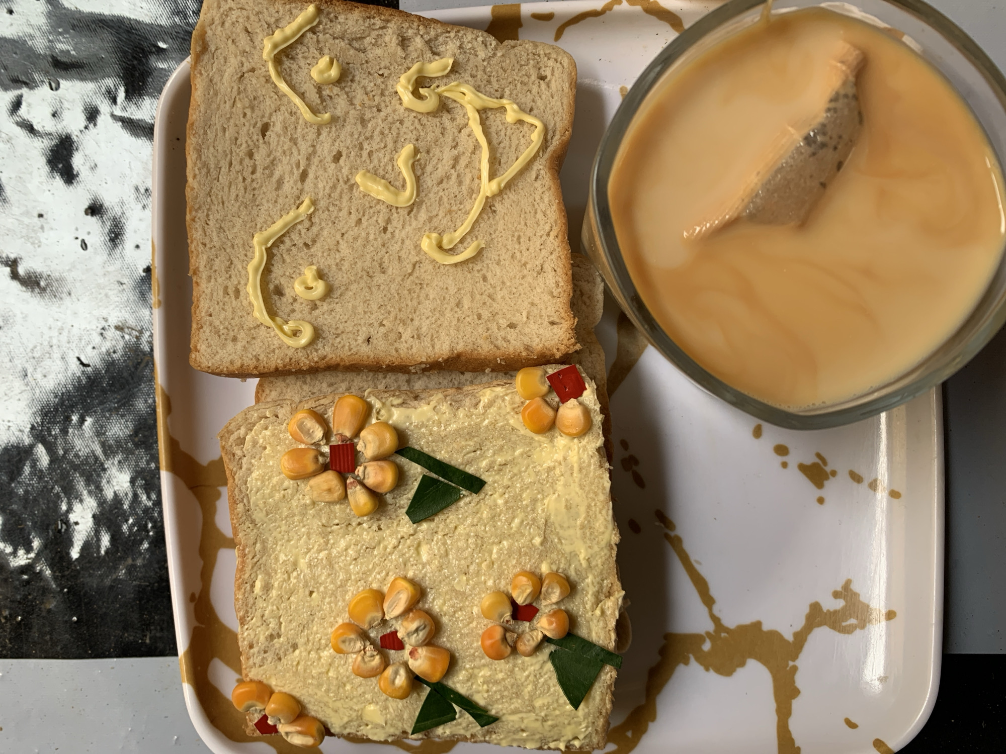 A white, rectangular tray holds two slices of bread. The top slice is decorated with lines and dots of yellow cream. The bottom slice is covered with yellow cream and decorated with corn kernels, diced red peppers, and green leaves, arranged to resemble flowers. To the right of the bread is a clear glass mug filled with a light brown liquid. A tea bag is submerged in the liquid. The tray has a white base with a pattern of brown and gold splatter. The background consists of a black and white textured surface on the left and a dark gray surface on the right.