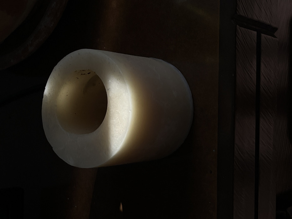 The image features a solitary, cylindrical object, likely a solid tube or a hollowed-out block, predominantly off-white or cream in color. The object is oriented horizontally, with its circular opening facing towards the left of the frame. It appears to be made of a dense, opaque material, possibly wax, plastic, or ceramic, and has a slightly textured surface with minor imperfections or dust specks visible, particularly on the inner rim and walls of the hollow center.
A strong, directional light source illuminates the object from the left, casting a bright highlight on the left-facing edge and the interior surface of the cylinder. This lighting creates a dramatic contrast, with the left side of the object brightly lit and gradually fading into shadow towards the right side. The right end of the cylinder is largely obscured by shadow.
The background is very dark and mostly indistinct, emphasizing the illuminated object. To the far right, there are glimpses of dark, vertical elements, possibly parts of a wooden structure or furniture, exhibiting some subtle texture. The surface beneath or behind the object is also dark, blending into the background. There is no visible text or discernable activity. The overall impression is one of a focused, artistic study of form and light on a simple, everyday object within a dimly lit environment.
