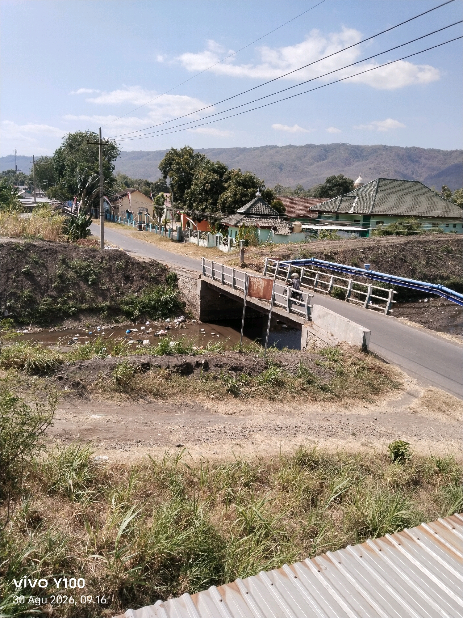 In Kendit, Indonesia, a rural scene unfolds under a clear blue sky with scattered clouds. A paved road runs through the frame, crossing a small, muddy river via a concrete bridge. The riverbed is littered with debris, including plastic bottles and wrappers, suggesting pollution. On either side of the road, dry, grassy embankments rise. Beyond the road, a cluster of houses with colorful facades and tiled roofs are visible, nestled amongst trees. One house features a green tiled roof and a white dome, possibly a mosque or religious building. Power lines crisscross the sky above the scene, and a lone figure on a motorcycle travels across the bridge. The foreground is dominated by dry grass and the corrugated metal roof of a structure. The timestamp "30 Agu 2026, 09.16" is overlaid on the bottom left of the image.