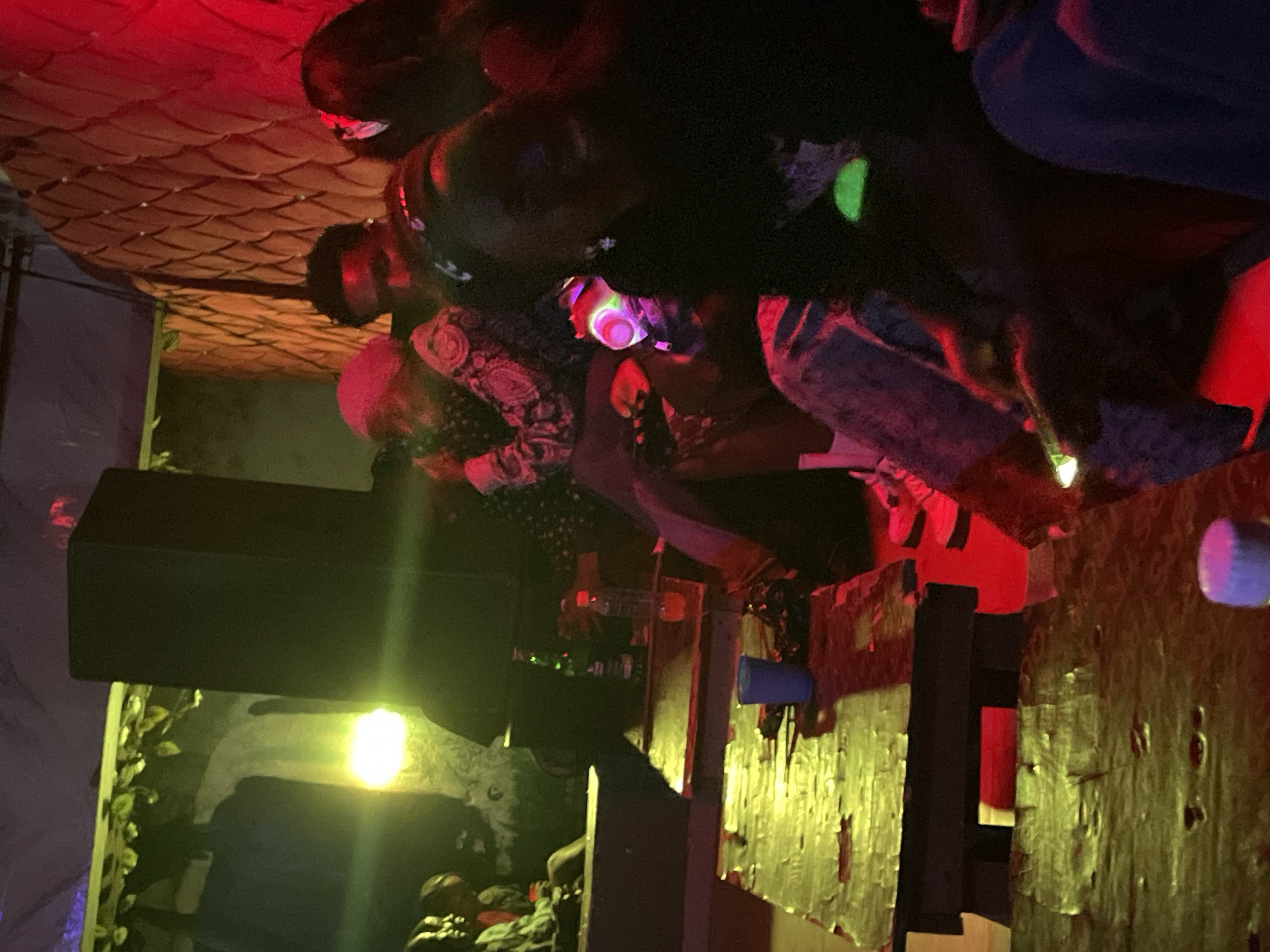 The image depicts a dimly lit indoor gathering, likely a club or bar, with predominantly red and green lighting. In the foreground, a wooden table with a worn, textured surface is visible, reflecting the colored lights. On the table, a blue plastic cup and a dark, crumpled object are present.
Further back, a large black speaker stands prominently. Several individuals are seated and standing around tables. One person, wearing a pink headscarf and a patterned top, is in the mid-ground. To their right, a man with dark hair appears to be looking down.
In the right mid-ground, a person wearing a black top is holding a glowing object, possibly a cup or bottle with a light inside, which emits a bright green and pink luminescence. Other individuals are partially visible in the background, suggesting a social gathering. The environment features a textured wall and some greenery with lights in the upper left quadrant. The overall atmosphere suggests a nightlife scene. The specific location in Durumi, Nigeria, cannot be determined from the image alone.