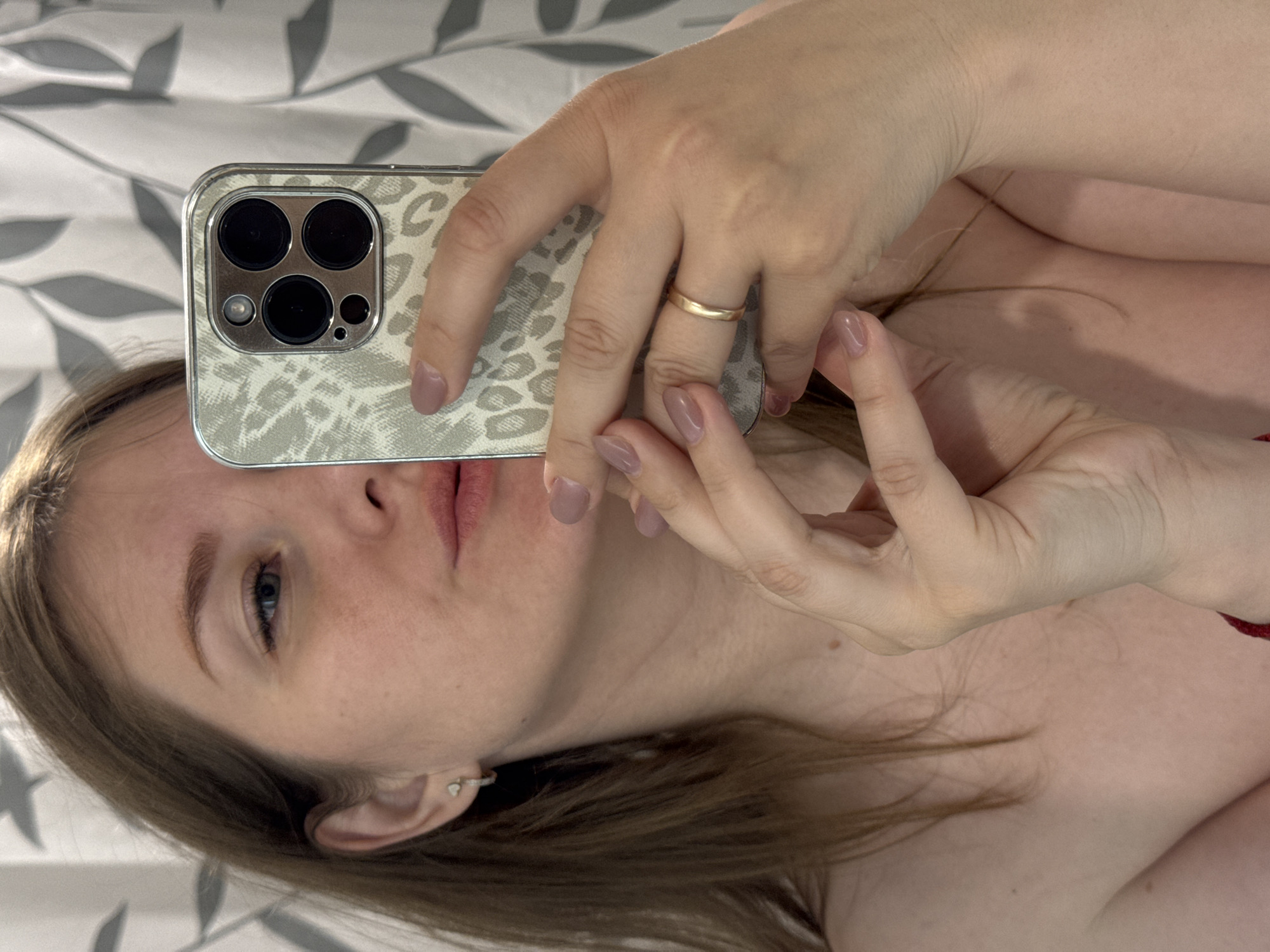 The image, oriented sideways, captures a person taking a selfie with an iPhone. The subject is a light-skinned individual, seen from the chest up, with light brown hair and blue/grey eyes. Their face is partially obscured by the smartphone, which they hold up with both hands.
The iPhone, likely a Pro Max model due to its three prominent camera lenses, is encased in a light-colored, possibly leopard print, cover. A simple gold-colored band ring adorns the person's left ring finger, and their fingernails are manicured with a light purple-taupe polish. A subtle earring is visible in their right ear. The subject's expression appears neutral or contemplative.
The background features a soft-patterned fabric, possibly bedding or a blanket, with a light grey and white leaf or organic design, indicating an indoor setting. The lighting is soft and diffused. No text is visible in the image.