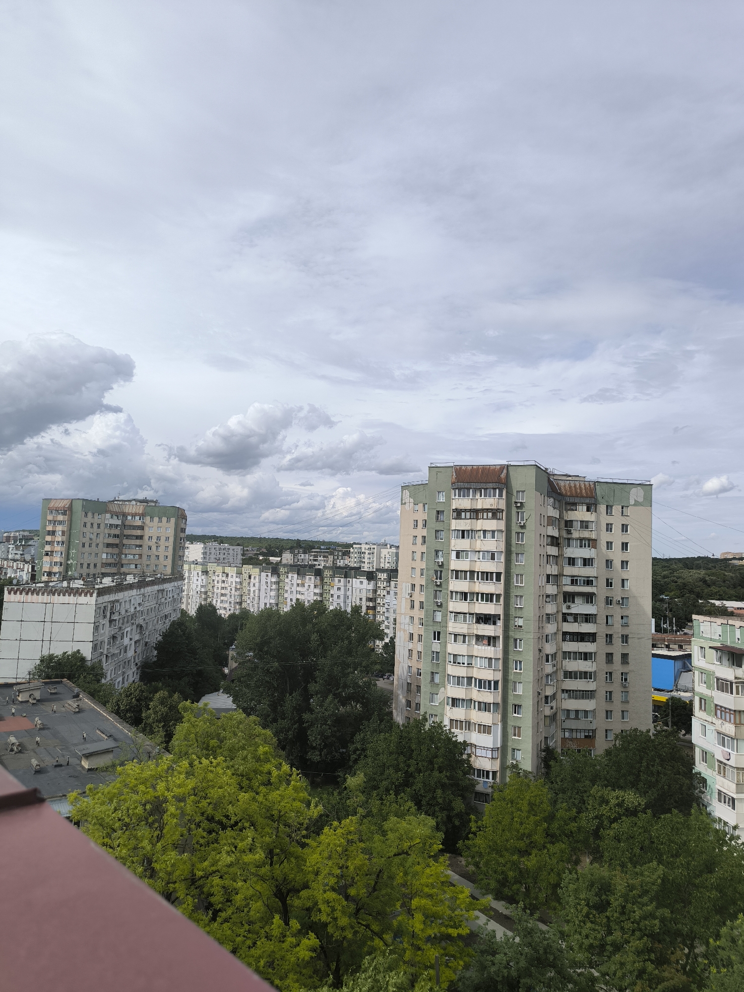The image depicts an urban landscape under a cloudy sky, likely taken from a high vantage point such as an apartment balcony. The scene is dominated by several multi-story apartment buildings, characteristic of Soviet-era architecture, with their uniform balconies and window arrangements. Some buildings are light-colored, while others have a pale green facade. Trees with lush green foliage are interspersed between the buildings, adding a touch of nature to the concrete environment.
The sky is filled with heavy, gray clouds, suggesting overcast weather, possibly before or after a rain shower. The lighting is diffused, indicating it is likely daytime, though the specific time is hard to determine due to the cloud cover. There are no visible people in the image.
Notable details include the weathered appearance of some of the apartment blocks, with some balconies showing signs of age and some rooftops displaying industrial-looking ventilation units. The presence of numerous electrical wires strung between buildings is also a common characteristic of urban infrastructure in many regions. In the background, a dense line of trees marks the edge of the urban area, hinting at a natural landscape beyond the city. One building in the distance has a bright blue and yellow structure adjacent to it, possibly a piece of street art or a temporary structure. There is no visible text in the image. The overall impression is that of a typical residential district in a city, captured on a cloudy day.