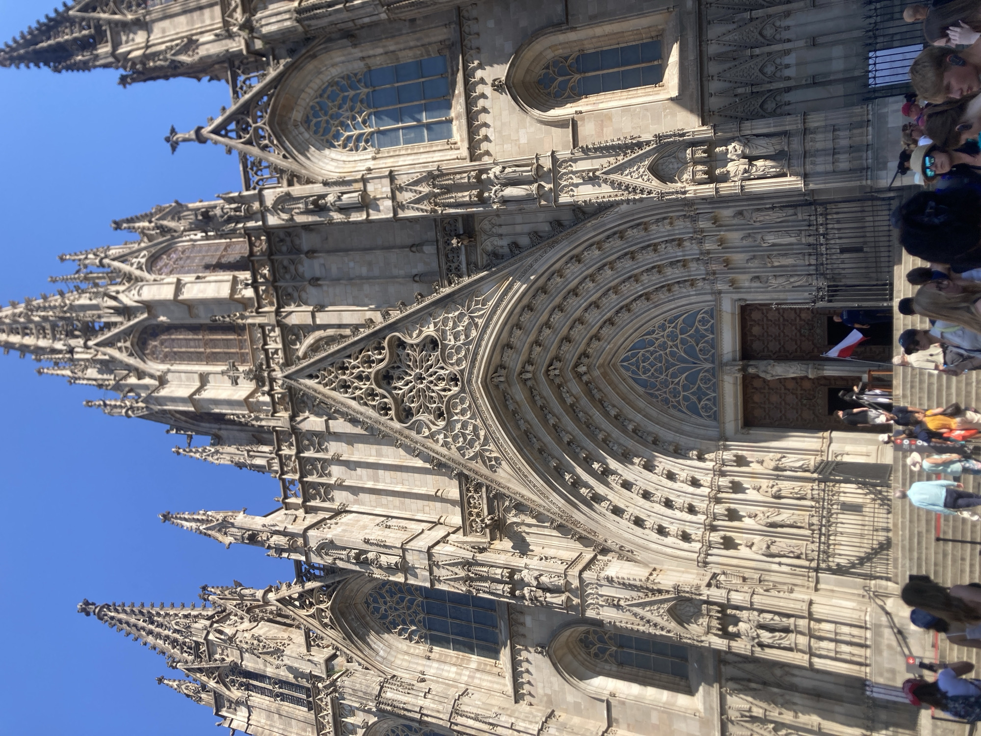 The image shows the facade of the Barcelona Cathedral, officially the Cathedral of the Holy Cross and Saint Eulalia, located in the Gothic Quarter of Barcelona, Spain. The cathedral's exterior is made of light-colored stone and features intricate Gothic architectural elements, including spires, pointed arches, gargoyles, and ornate carvings. Tall, slender windows with tracery are visible. The sky above is a clear, bright blue, indicating a sunny day.
In the foreground, a group of tourists are gathered on the steps leading up to the cathedral entrance. They are dressed in casual summer clothing. Some are looking at the cathedral, while others appear to be talking or taking pictures. One person near the entrance is holding a Polish flag. Several people are partially obscured by the foreground, but their presence indicates a public gathering. The scene is a typical tourist snapshot of a famous landmark.