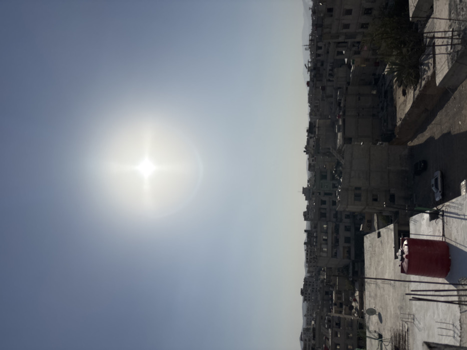 The image displays an elevated, wide shot of a dense urban environment in Mu'adammiyat ash-Sham, Syria, under a clear, bright sky. A prominent sunburst, indicative of direct sunlight, is visible in the upper-center of the frame.
The cityscape consists primarily of multi-story buildings constructed from concrete and masonry, appearing in shades of gray, beige, and light brown. Many structures exhibit visible signs of damage, decay, or incomplete construction, with exposed rebar protruding from rooftops and walls, and missing window sections. Buildings are densely packed, forming narrow gaps and unpaved pathways at ground level.
In the lower-left foreground, a concrete rooftop features a red, cylindrical water tank. To its left, another concrete rooftop has a white satellite dish and several vertical rebar elements. Below these rooftops, a dirt path or unpaved road shows two parked vehicles: a light-colored sedan (likely white) in the foreground and a darker vehicle behind it. Sparse, green vegetation is visible between some buildings towards the right. The distant horizon reveals faint, hazy hills or mountains. No individuals are depicted, and no specific actions or interactions are discernible.