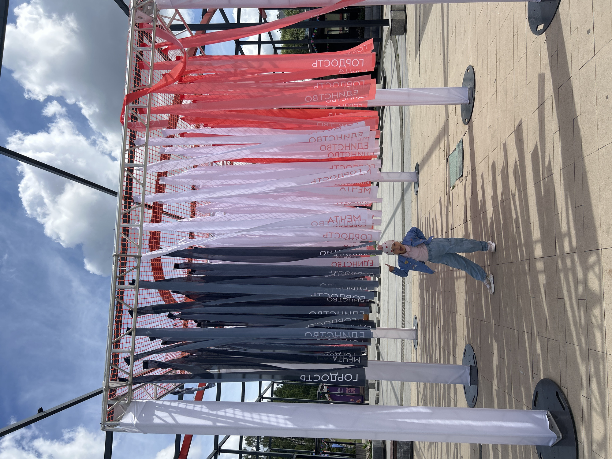A young girl, dressed in a denim jacket, pink shirt, and blue cargo pants, stands in front of a display structure. She is pointing upwards with her right index finger and has her mouth open, seemingly shouting or exclaiming. The display structure consists of vertical white fabric panels on the sides and a metal frame overhead, from which numerous vertical fabric ribbons of varying colors - dark gray, white, and red - are hanging. Some of the ribbons have text printed on them in white lettering. The ground is paved with light-colored square tiles. In the background, there are trees and a blue sky with scattered white clouds. The scene appears to be outdoors in a public area. The text visible on the ribbons, such as "ГОРДОСТЬ" (Pride), "МЕЧТА" (Dream), and "ЕДИНСТВО" (Unity), suggests this might be part of a cultural or patriotic exhibition. This location is likely within Izhevsk, Russia, given the Cyrillic text.