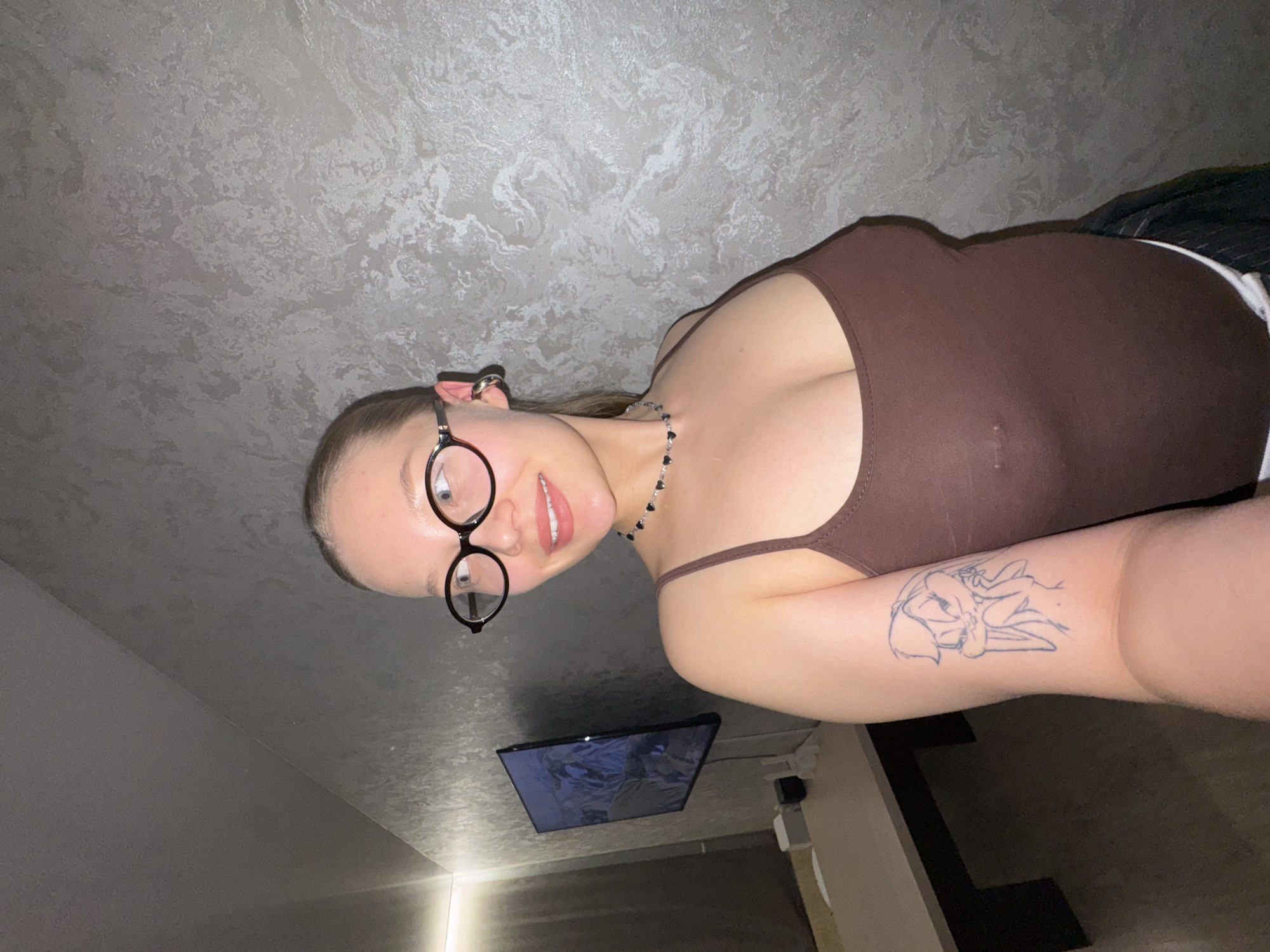 A woman is shown indoors with a textured grey wall and ceiling in the background. She is wearing a brown tank top, a black necklace with heart-shaped beads, and round-framed glasses. She has a tattoo on her upper arm. A television is visible in the background, along with what appears to be a cabinet. The location is Chaikovsky, Russia.