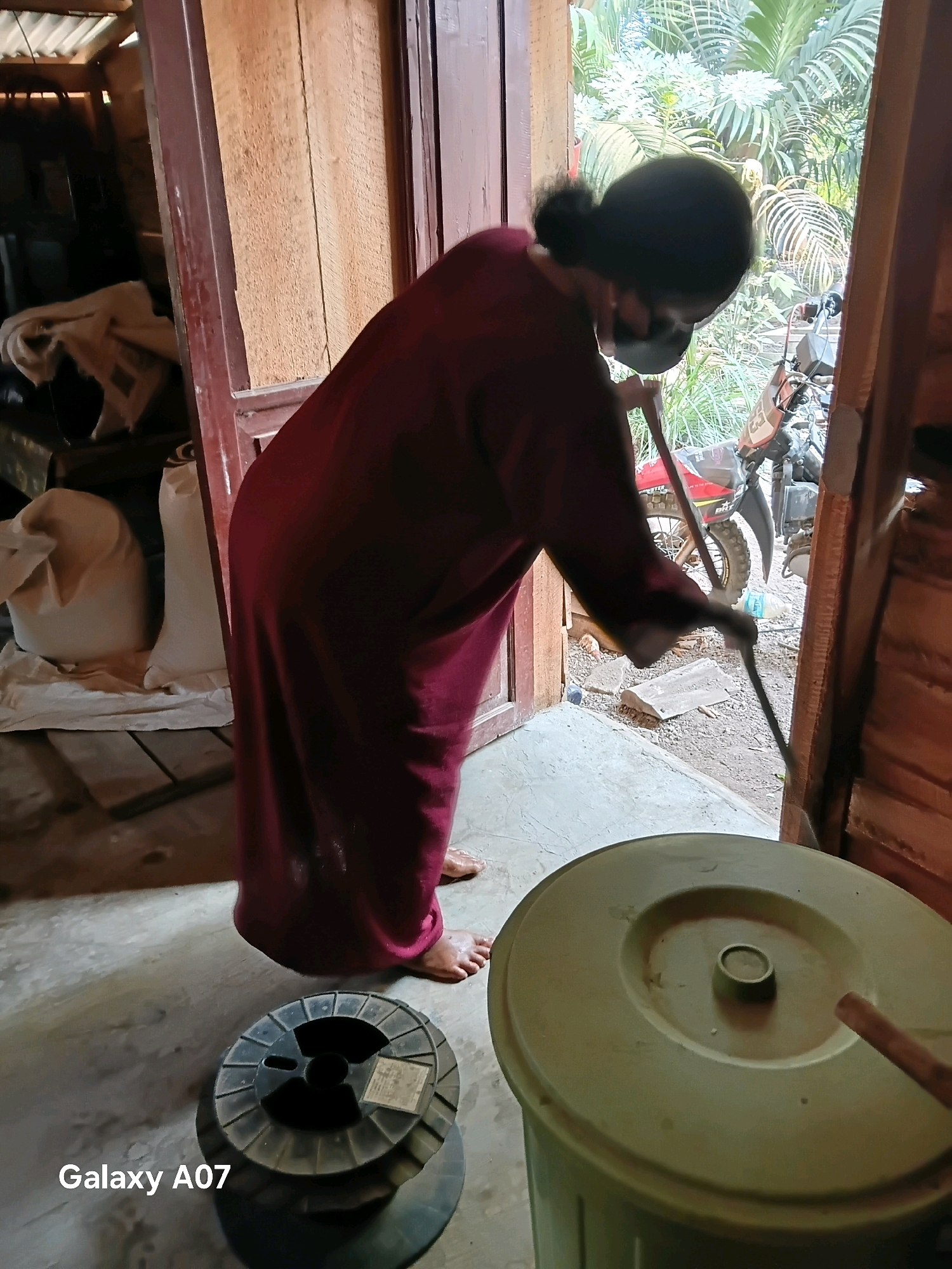 A person wearing a maroon robe and a black face mask is sweeping the floor with a broom. The person is standing in what appears to be an open doorway, with a motorcycle visible in the background outside. Inside the doorway, there are large sacks of what appears to be grain. In the foreground, there is a large green trash can and a spool of wire. The scene is likely a residence or storage area in Margo Rukun, Indonesia.