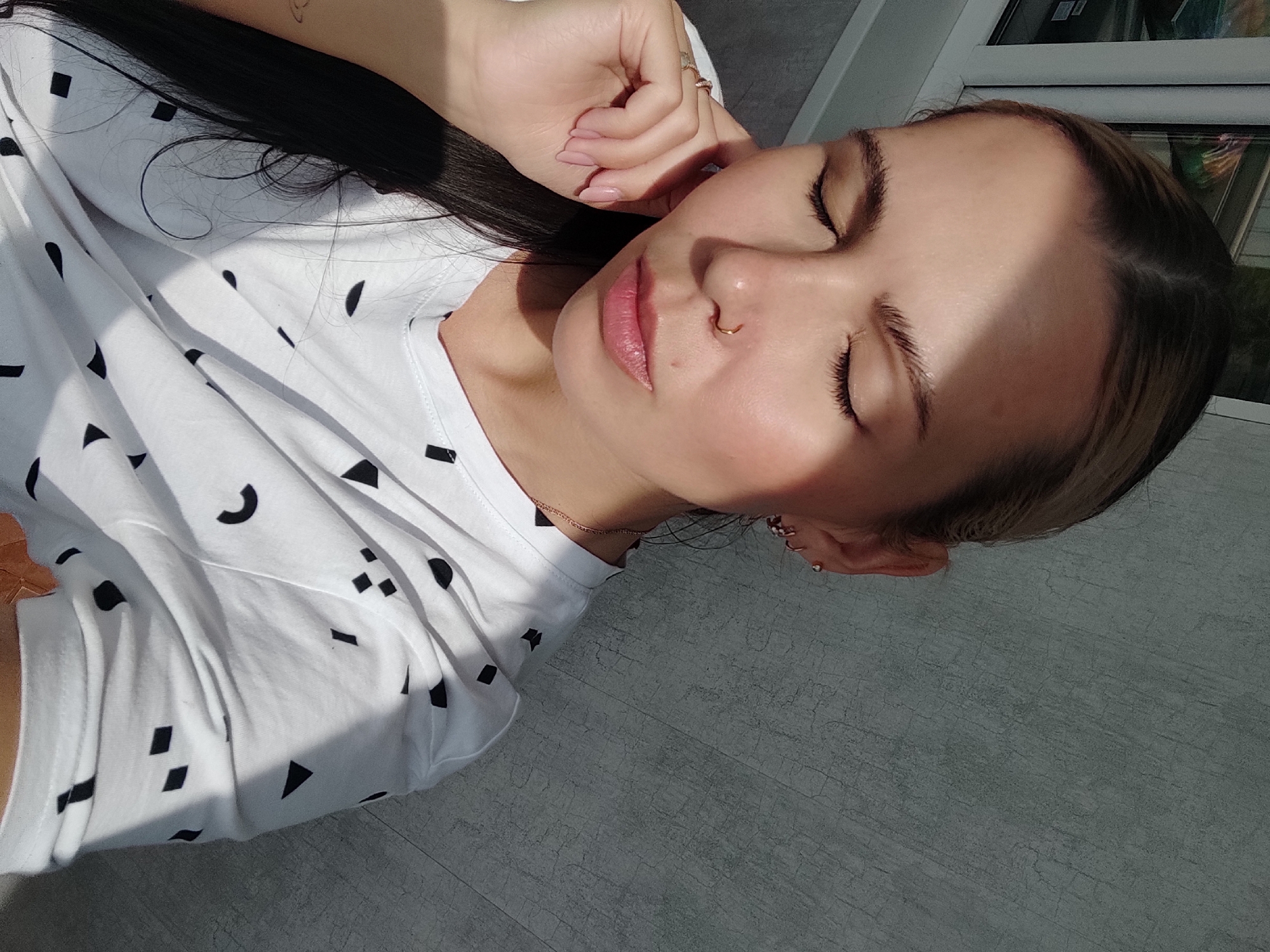 A young woman is lying down indoors on a gray tiled floor. She has fair skin and brown hair with blonde highlights pulled back from her face. She is wearing a white t-shirt with small black shapes printed on it. Her eyes are closed and she is facing the camera. She has a nose ring in her left nostril. Her left hand is touching her face. There is a window in the background. The photograph was taken in Chaikovsky, Russia.