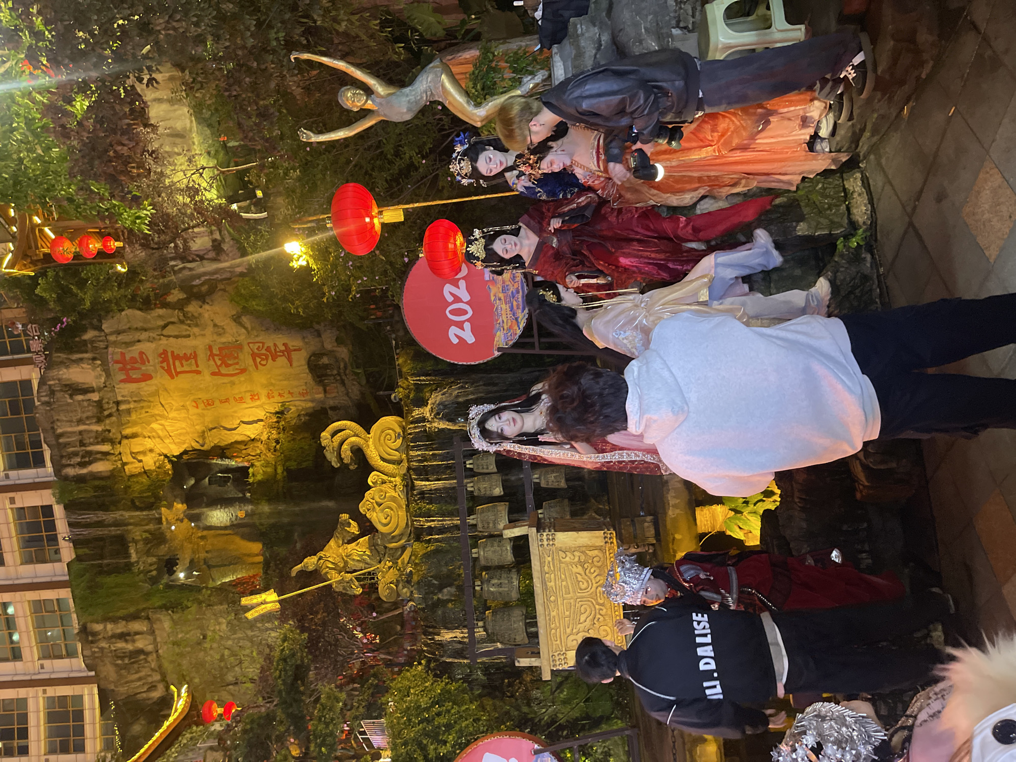 The image depicts a nighttime cultural event or photoshoot taking place in Yuzhong District, China. The scene is illuminated by artificial lights, including red lanterns and yellow spotlights, against a prominent, multi-tiered rock face. A waterfall cascades down the rock face, which also features traditional Chinese characters inscribed vertically and a large golden statue of a robed figure holding a staff with banners, situated above a multi-headed golden serpent sculpture. Below this, a set of bronze bells is arranged on a decorative stand. At the top of the rock face, a modern building with multiple windows is visible.
In the foreground and midground, a group of individuals is present. Five to six people are dressed in elaborate traditional Chinese costumes, featuring intricate headpieces, rich fabrics in shades of red, gold, blue, and orange, and detailed makeup. They appear to be posing for photographs. One woman in a red and gold costume with a white feathered headpiece stands centrally, while others are arranged around her, some seated on ledges. To the right, a golden statue of a slender figure with arms raised in a dance pose is visible. A large red circular sign displaying "2023" is positioned behind some of the costumed figures.
Several individuals are involved in photography or observation. One man, seen from the back, wears a light grey hoodie and dark pants, holding a mobile phone towards the scene. Another man, in a black leather jacket and dark pants, actively operates a professional camera with an attached flash, directing his lens towards the costumed subjects. A third man, viewed from the back, wears a black jacket with "LI. DALISE" printed on it and dark pants, interacting with one of the costumed individuals. Another person in a silver intricate headpiece and red costume is visible on the far left. The ground consists of patterned paving stones. A white plastic stool is visible near the photographer on the right. The overall environment suggests a festive or themed cultural attraction.