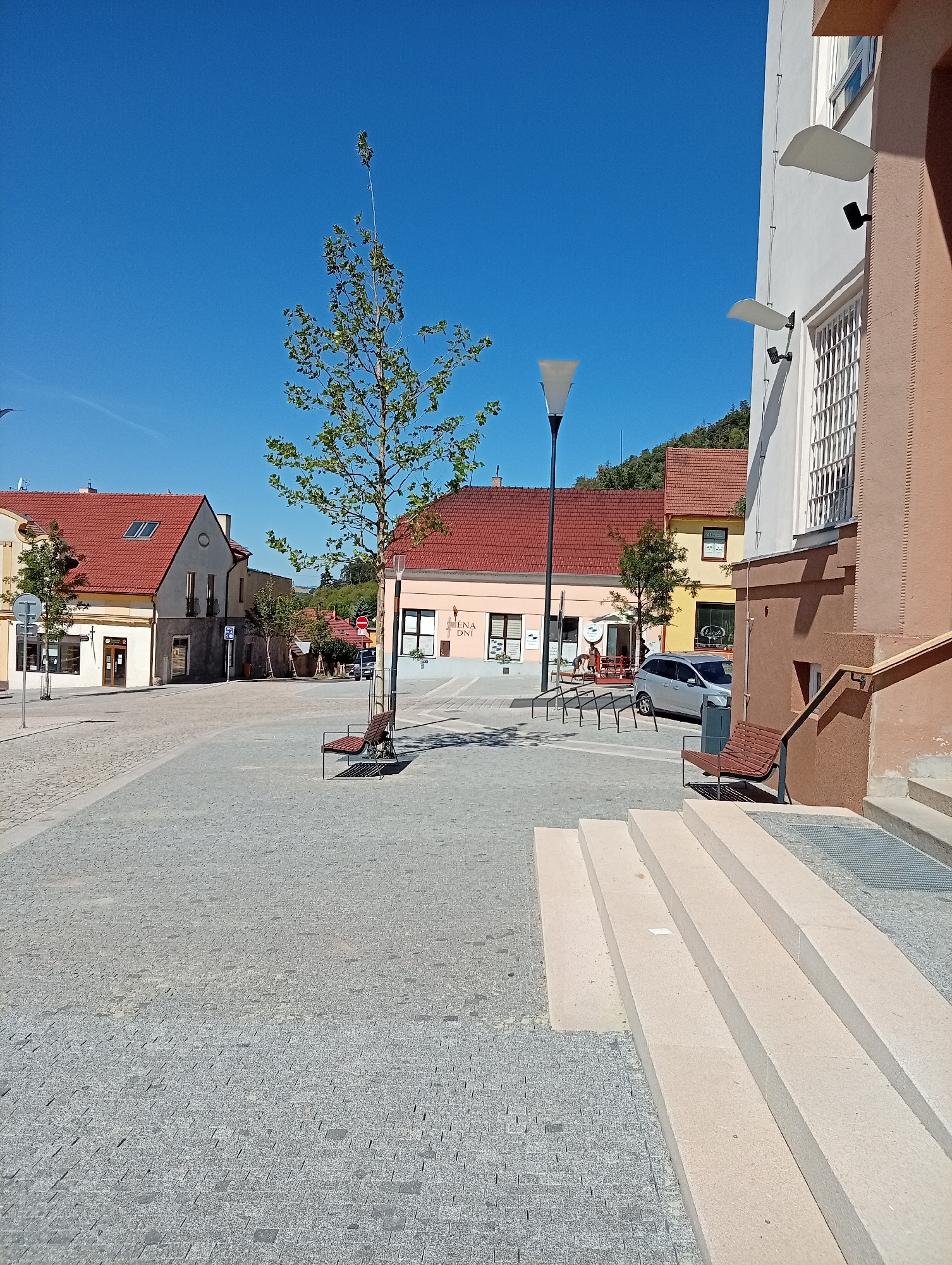 The image depicts a street scene in Tišnov, Czechia, specifically on Denisova Street, near the post office. A modern building with light brown and white facades is prominently featured on the right side of the frame, featuring a large barred window and a staircase with railings. Adjacent to this, a street lamp with a triangular shade stands tall. The street itself is paved with cobblestones and features several benches. Across the street, a row of older buildings with red tiled roofs line the square. One building has the signage "NABÍDKA PRO DNEŠEK" and what appears to be a cafe or shopfront with tables and chairs outside. A silver car is parked on the street. The scene is bathed in bright sunlight, casting shadows on the ground. The sky is clear and blue, indicating a sunny day. A young tree with sparse foliage is planted in the middle of the square.