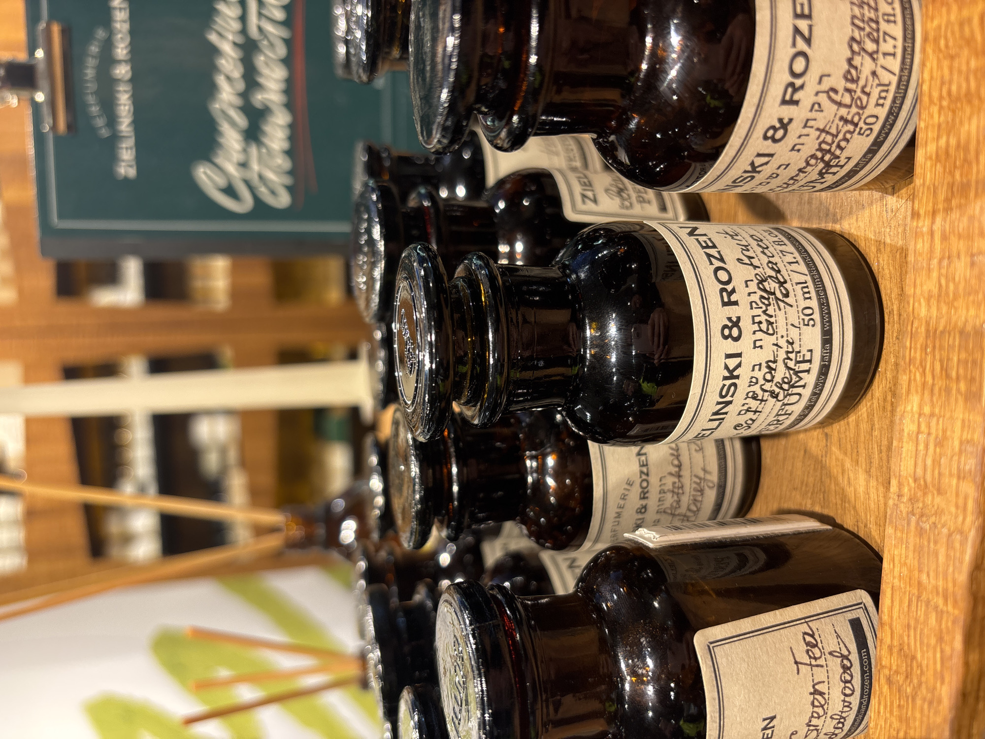 The image shows a close-up view of several small, dark amber-colored glass bottles with cylindrical bodies and clear glass stoppers. They are arranged in rows on a light-colored wooden surface. Each bottle has a beige label with black text, which includes the brand name "Zielinski & Rozen". The labels also feature ingredient information such as "Grapefruit & Tobacco", "Honey", "Green Tea & Salwood", and "Amber & Leather". Text on the labels appears to be in both English and Hebrew. The bottles contain what appears to be perfume or scented oils, with some having thin wooden reeds inserted.
In the background, out of focus, there are more rows of bottles and what appears to be a green sign with white and red lettering. The sign reads "Gourmet & Authentic".
The location is not specified in the image description.