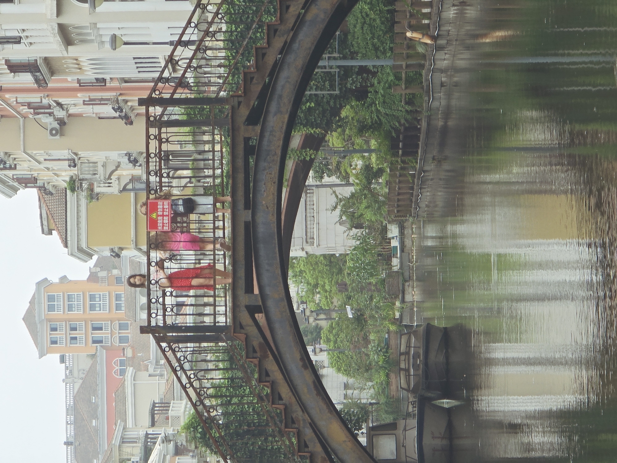 The image depicts a canal in the city of Xiaoshan District, China, with a pedestrian bridge spanning it. Two young women are standing on the bridge, one wearing a red dress and the other a pink dress. A red sign with Chinese characters and a warning symbol is visible on the railing near the women. The canal is flanked by buildings with architectural styles reminiscent of European towns, featuring pastel colored facades and balconies. Two boats are docked on the left side of the canal, partially submerged in the water. The water reflects the buildings and greenery, creating a serene, albeit somewhat overcast, atmosphere. Lush vegetation lines the canal banks, with trees and plants visible behind the railing of the bridge and along the water's edge. The scene suggests a picturesque urban waterway, likely a tourist or residential area.