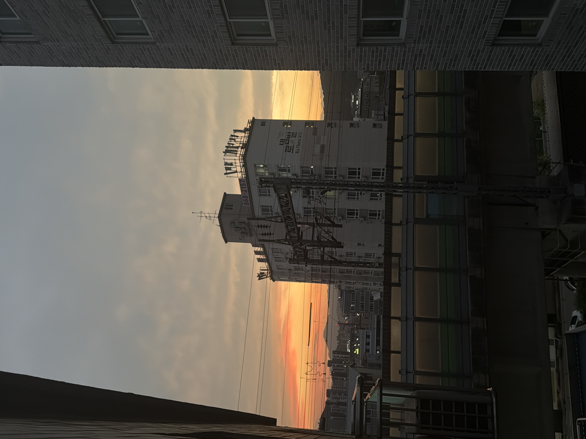 The image depicts an urban cityscape in Dongducheon-si, South Korea, at dusk. A prominent multi-story building with visible windows and antennae on its roof stands in the center, bearing the Korean text "더 로 뎀발" and "RoThem Vill". Electrical infrastructure, likely for a railway, runs across the foreground and midground. In the background, a range of hills is visible against a sky filled with the warm hues of sunset. Residential buildings and streetlights are scattered throughout the city. The foreground is partially obscured by the dark silhouette of a building on the left and the textured brick facade of a building on the right. A walkway or elevated structure with glass panels runs horizontally across the midground.
