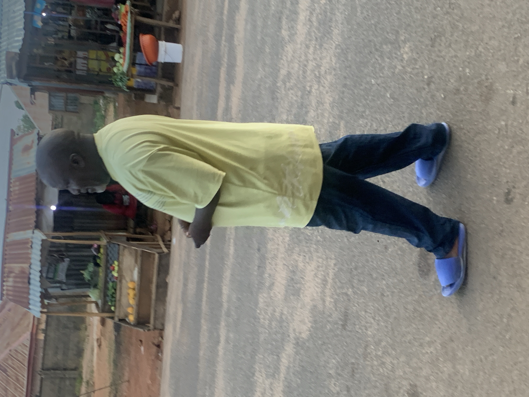 The image depicts an outdoor street scene, likely in Barakin Akawo, Nigeria. The primary subject is a bald, dark-skinned man standing on a grey, paved surface. He is dressed in a light yellow short-sleeved shirt, dark blue jeans, and light blue slide sandals. The man is oriented with his right side facing the viewer, his head tilted downwards and to his left, suggesting he is looking at something on the ground or just out of frame. His hands appear to be clasped together in front of his waist.
In the background, to the left of the man, are several market stalls constructed from wooden frames and corrugated metal roofs. These stalls display various items, including what appears to be fresh produce such as green vegetables and yellow fruits. An orange basket and a white bucket are visible near one of the stalls. A small, indistinct portion of what might be another person or an object in red is partially visible behind the man's lower left side.
The scene is set during the daytime, with natural, somewhat diffused light, possibly indicating an overcast sky or early morning/late afternoon. The general environment suggests a bustling local market or roadside commercial area. No specific activity is captured beyond the man standing, and no visible text is present.