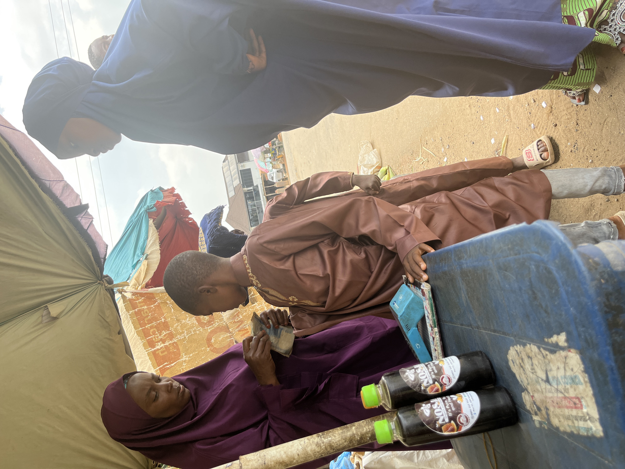 This media file depicts an outdoor scene, likely a market or roadside stall, in Abuja, Nigeria. The central subjects are two individuals, one lying down and the other seated. The person lying down is wearing a deep purple hijab and garment, and is holding a wad of Nigerian Naira currency, appearing to be counting it. The seated individual, dressed in a brown kaftan, is looking down at the money.
The setting includes a makeshift tent or shelter made of tarpaulins in various colors, suggesting a temporary market stall. In the foreground, a blue plastic container is visible, adorned with stickers, one of which appears to be a worn-out label with some visible text. Two dark brown bottles with green caps are placed on the container; their labels are partially visible and read "NOSI NOS," with further text indicating "Organic and Pure," and "It's Pure, and Hea... and h...". A small blue electronic device, possibly a calculator or payment terminal, is also present.
The ground appears to be sandy, with scattered debris. The lighting suggests it is daytime, possibly late morning or early afternoon, with bright sunlight casting shadows. The overall mood appears to be one of commerce and daily transaction. The visible currency and the presence of a market stall indicate a commercial activity taking place. The individuals' clothing suggests a cultural context common in Nigeria.