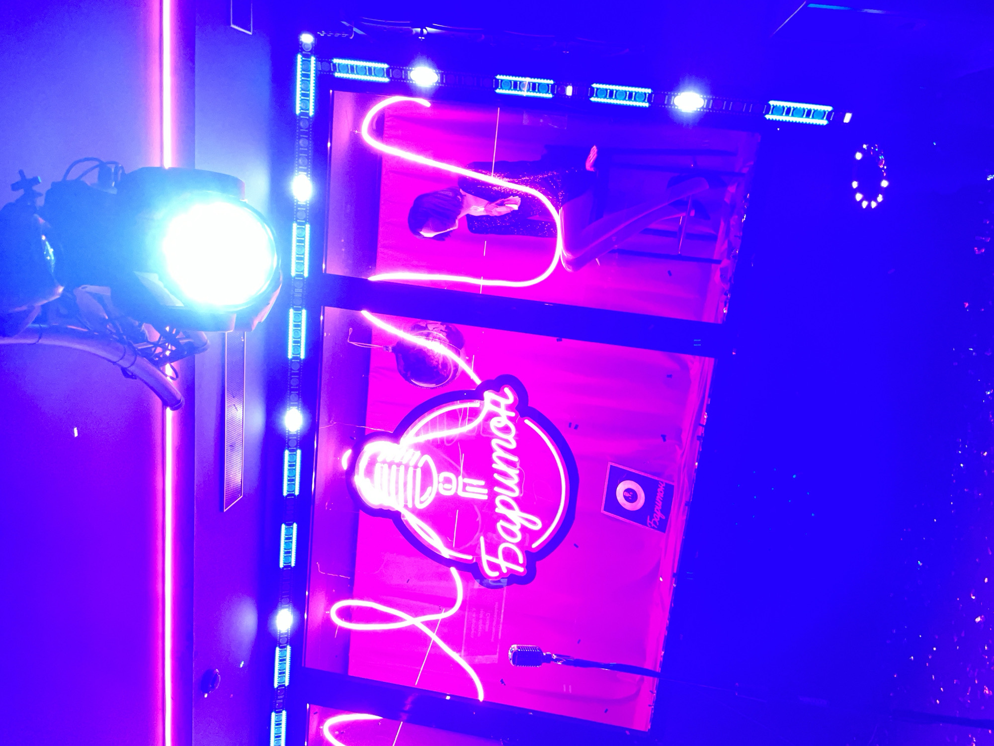 The image shows a neon sign in Perm, Russia. The sign features a microphone design enclosed in a circle with the word "Баритон" (Baritone) in Cyrillic script alongside a stage lit with neon lights. A person is visible in the top panel of the sign. A spotlight is positioned to the left of the sign.