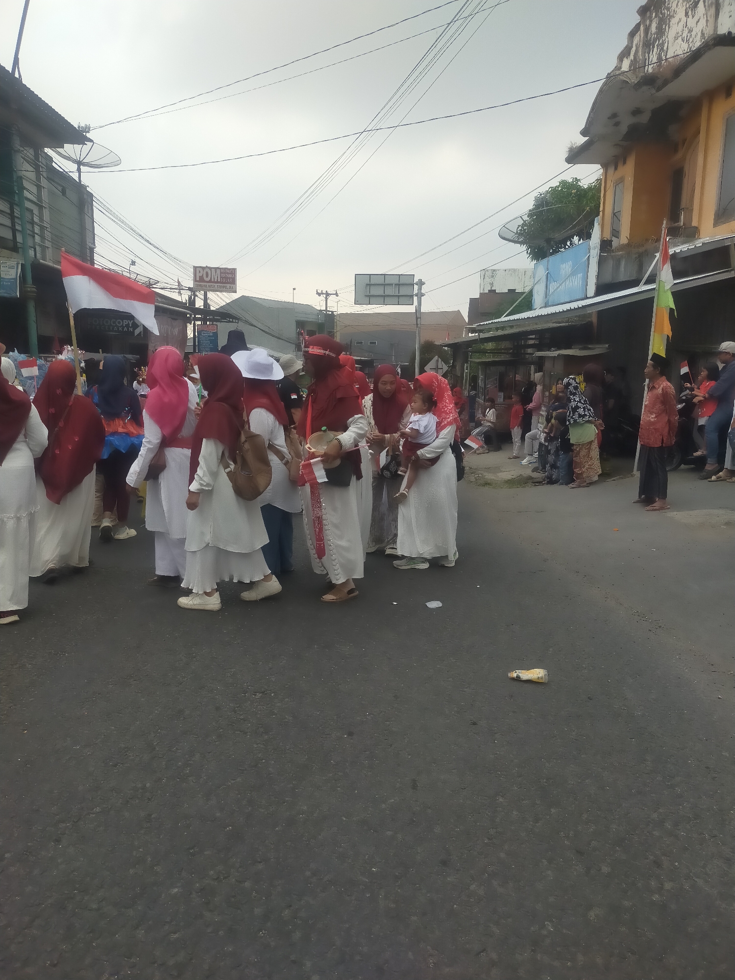 In the city of Tuwel, Indonesia, a group of people are gathered on a paved street under an overcast sky. Many individuals, predominantly women, are wearing white or red traditional clothing, with some also wearing headscarves. A few are holding small Indonesian flags, indicating a possible parade or celebration. In the background, buildings line the street, some with faded paint and satellite dishes. A sign with the text "POM" is visible on one of the buildings. The overall scene suggests a community event taking place on a relatively quiet street.
