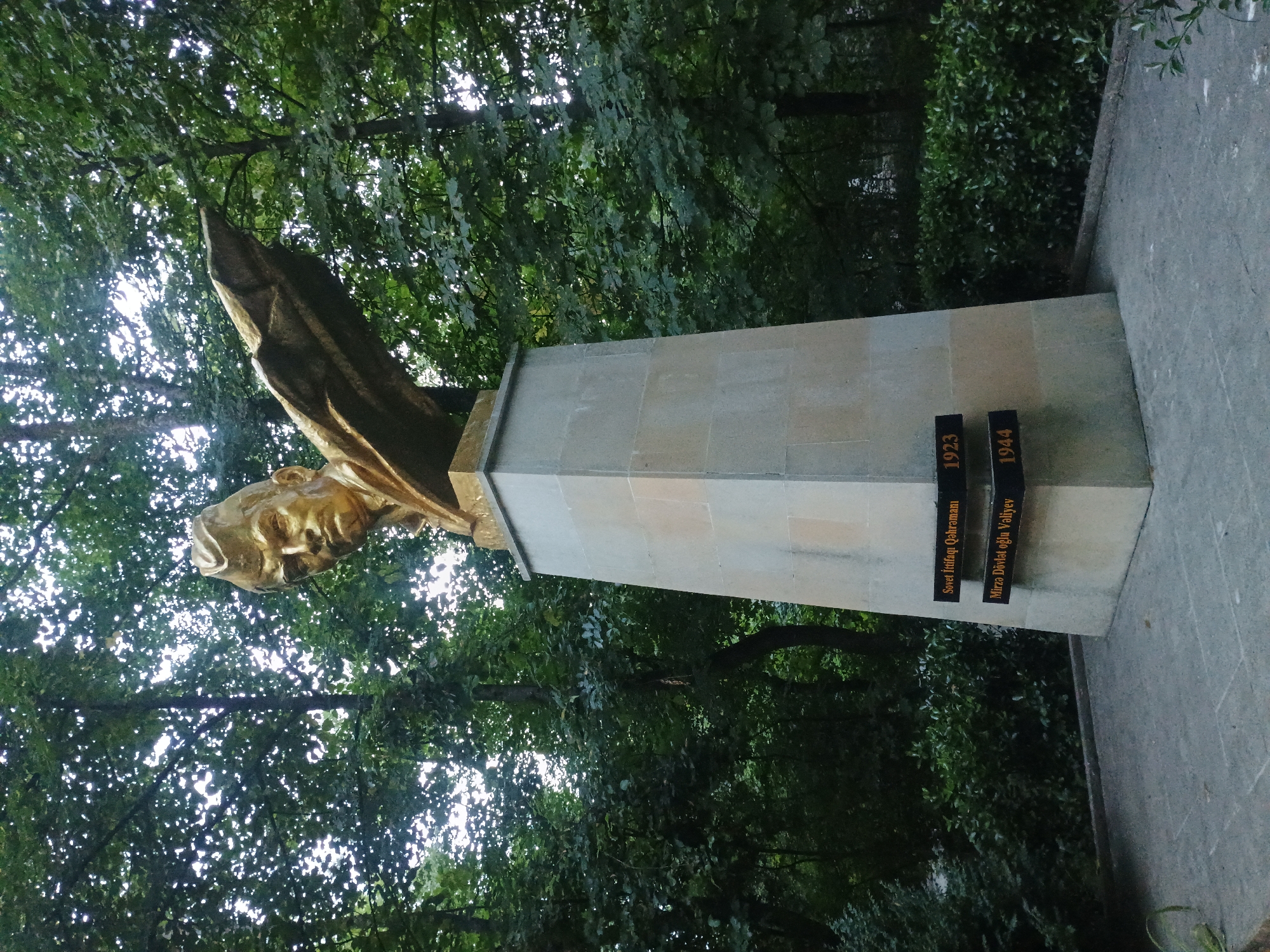 This is a full-shot, eye-level, outdoor photograph of a golden bust of a man wearing a military cap, mounted on a tall, light-colored stone pedestal. The bust faces slightly to the left, with its head turned to look directly at the camera. Its left arm is extended outwards, depicted as a wing-like structure. The pedestal is triangular in cross-section and has two black plaques on its front face. The top plaque reads "Sovet Ittifaqi Qəhrəmanı 1923" and the bottom plaque reads "Mirzə Dövlət oğlu Vəliyev 1944". The monument is situated in a park-like setting with lush green trees and bushes in the background. The ground in front of the monument is paved with grey stones. The lighting suggests it is daytime, possibly late afternoon, with dappled sunlight filtering through the trees. This is the monument to Hero of the Soviet Union, Mirzə Dövlət oğlu Vəliyev, located in the city of Qusar, Azerbaijan.