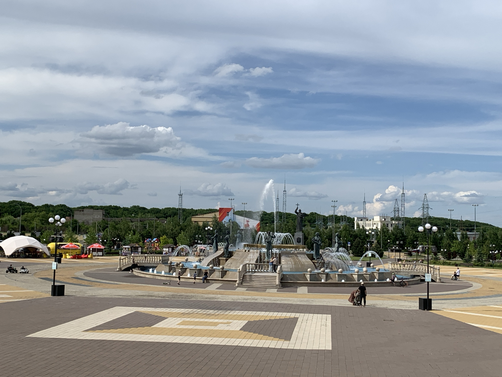 An outdoor, wide-angle, eye-level, day-time photograph captures a large, multi-tiered fountain adorned with sculptures and water jets, centrally located within a paved plaza in the city of Unknown, Unknown. The fountain is the primary subject, surrounded by a circular path and various attractions including a white tent with tables and chairs, a yellow food truck, and red umbrellas. Several people are visible in the scene; a person pushes a stroller on the right, another rides a bicycle, and a few individuals are observed near the fountain. The plaza is paved with brick and decorative patterns. In the background, a dense line of green trees forms a backdrop, behind which are buildings and electrical towers, suggesting an urban environment. The sky above is filled with scattered white clouds and patches of blue.