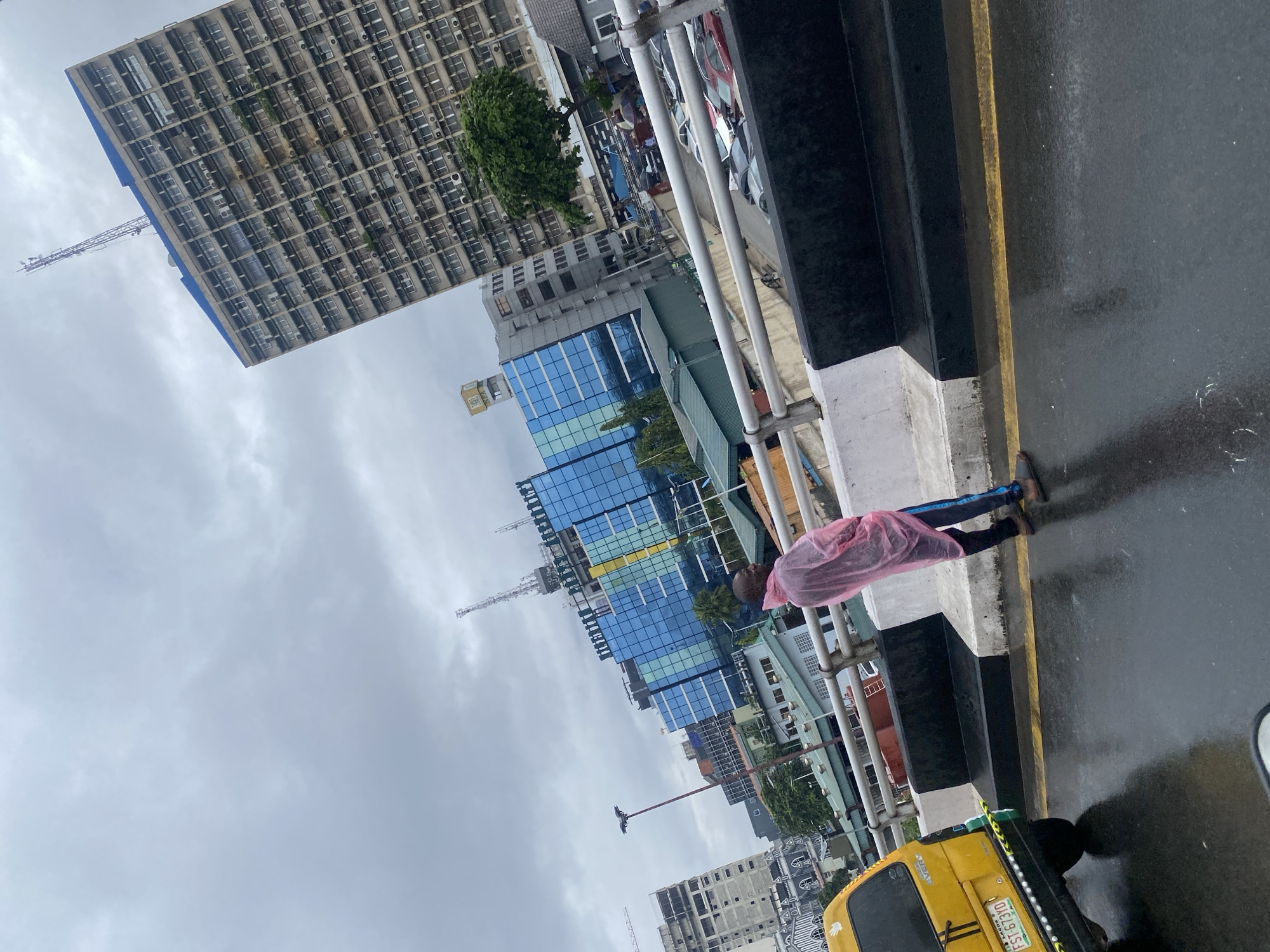 The image depicts a scene on a wet, possibly rain-slicked road, on Victoria Island, Nigeria. The perspective is from a vehicle, looking forward and slightly to the right. In the foreground, a yellow taxi with license plate "FST-673YD" is partially visible on the left. A person wearing a pink plastic poncho and dark pants stands on the side of the road, facing away from the camera and looking towards the cityscape.
The background features a cluster of modern buildings. Prominently, a tall, grey, brutalist-style building with numerous windows and air conditioning units dominates the right side of the frame, topped with a telecommunications mast. To its left, a more contemporary building with a blue, glass-paneled facade rises, featuring a prominent yellow sign with the letters "NTA" (Nigerian Television Authority). Other buildings of varying heights and architectural styles are visible, along with trees and what appear to be construction cranes in the distance. The sky is overcast and cloudy, contributing to the muted lighting of the scene. The wet road surface reflects the surrounding lights and buildings, indicating recent or ongoing rainfall.