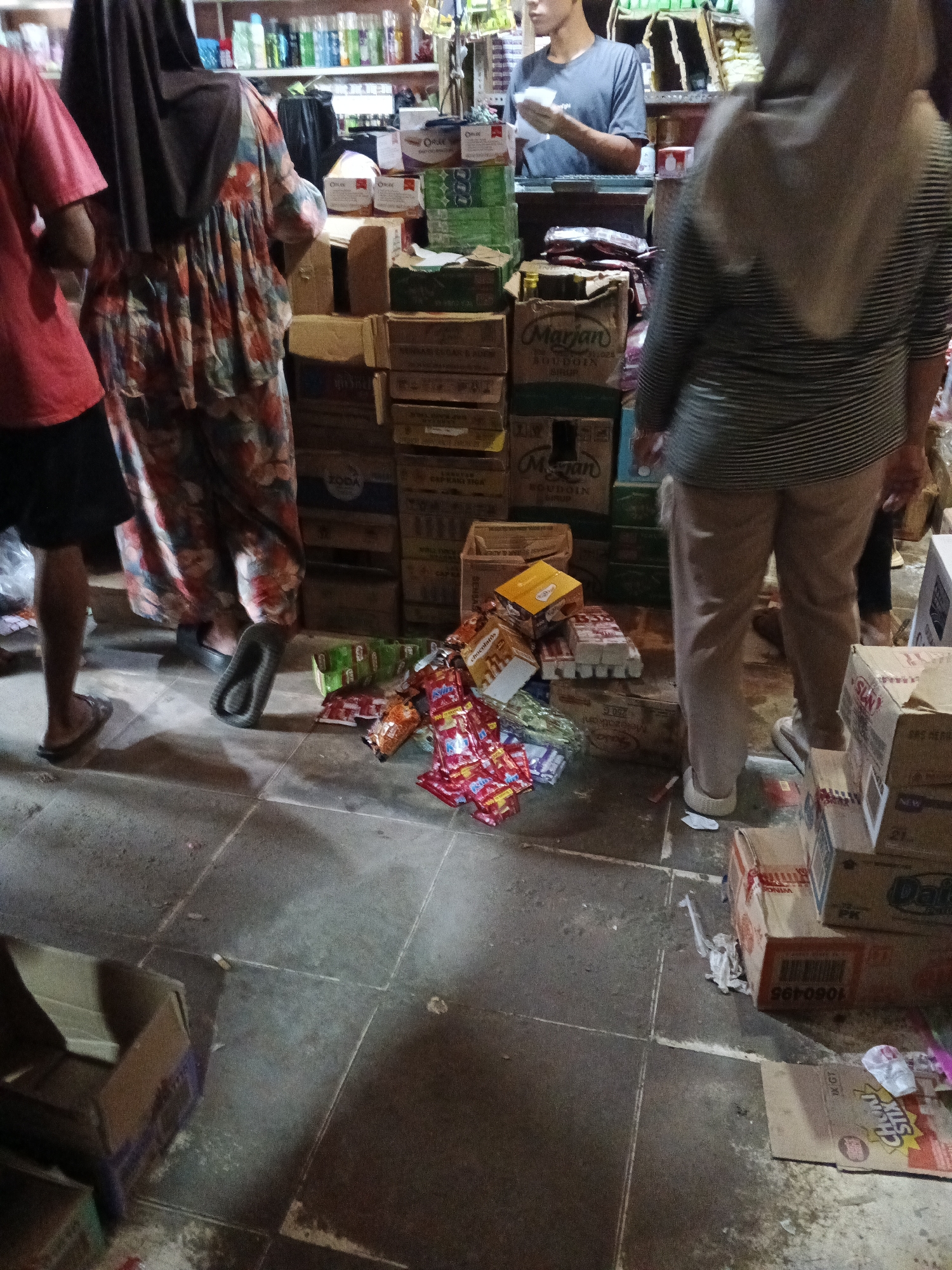 The image depicts an outdoor scene in what appears to be a market or store in Warungkiara, Indonesia. The foreground shows a tiled floor with several cardboard boxes stacked and scattered. Some boxes are open, revealing their contents, which include what looks like packaged snacks or food items. There are also loose wrappers and plastic debris on the floor.
In the mid-ground, three individuals are visible. On the left, a person wearing a red shirt and dark shorts is partially visible, facing away from the camera and appearing to be walking. In the center, a person wearing a floral dress and a dark headscarf is also facing away and walking. A pair of black flip-flops are visible near this person's feet. To the right, a person in a beige headscarf and a striped shirt, wearing beige pants and white slip-on shoes, is standing and looking towards the right. Behind this individual, another person in a grey t-shirt is standing and holding a white item, possibly a receipt or product.
The background reveals shelves stocked with various items, including bottles and boxes, suggesting a retail environment. The overall impression is that of a busy marketplace with goods being organized or sold.