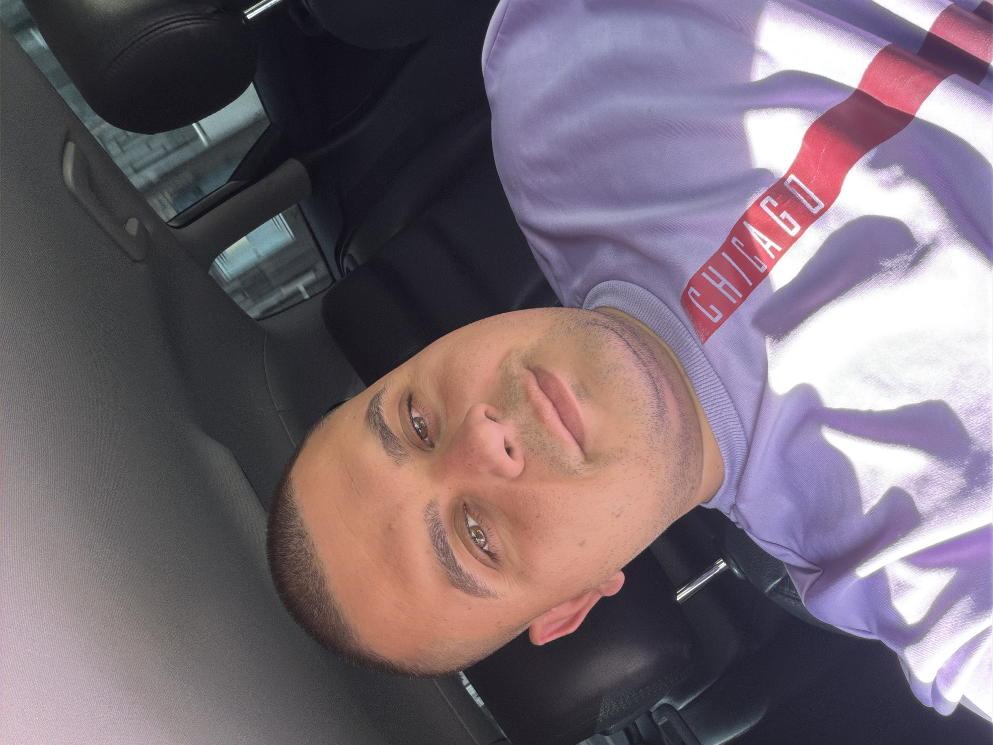 A young man is captured in a close-up, upward-angled shot, likely a selfie, inside what appears to be a car in Shumen, Bulgaria. He looks directly at the camera with a neutral to slightly contemplative expression, featuring a subtle smirk. He has short, dark, closely-cropped hair, light brown eyes, and stubble on his chin and jawline, with a faint, thin mark or scar visible on his left cheek/jawline.
He is wearing a light purple, long-sleeved sweatshirt. A prominent red band runs vertically across the front of the sweatshirt, featuring the word "CHICAGO" in white capital letters. Bright sunlight illuminates the scene, casting strong, distinct shadows on his face and across his sweatshirt, indicating that it is a sunny daytime.
The background shows elements of a vehicle's interior. Dark, possibly leather, car seats are visible, along with a lighter grey fabric on the ceiling or pillar where a grab handle is mounted. Reflections in the car's window or sunroof reveal what appears to be parts of a building's structure outside the vehicle.