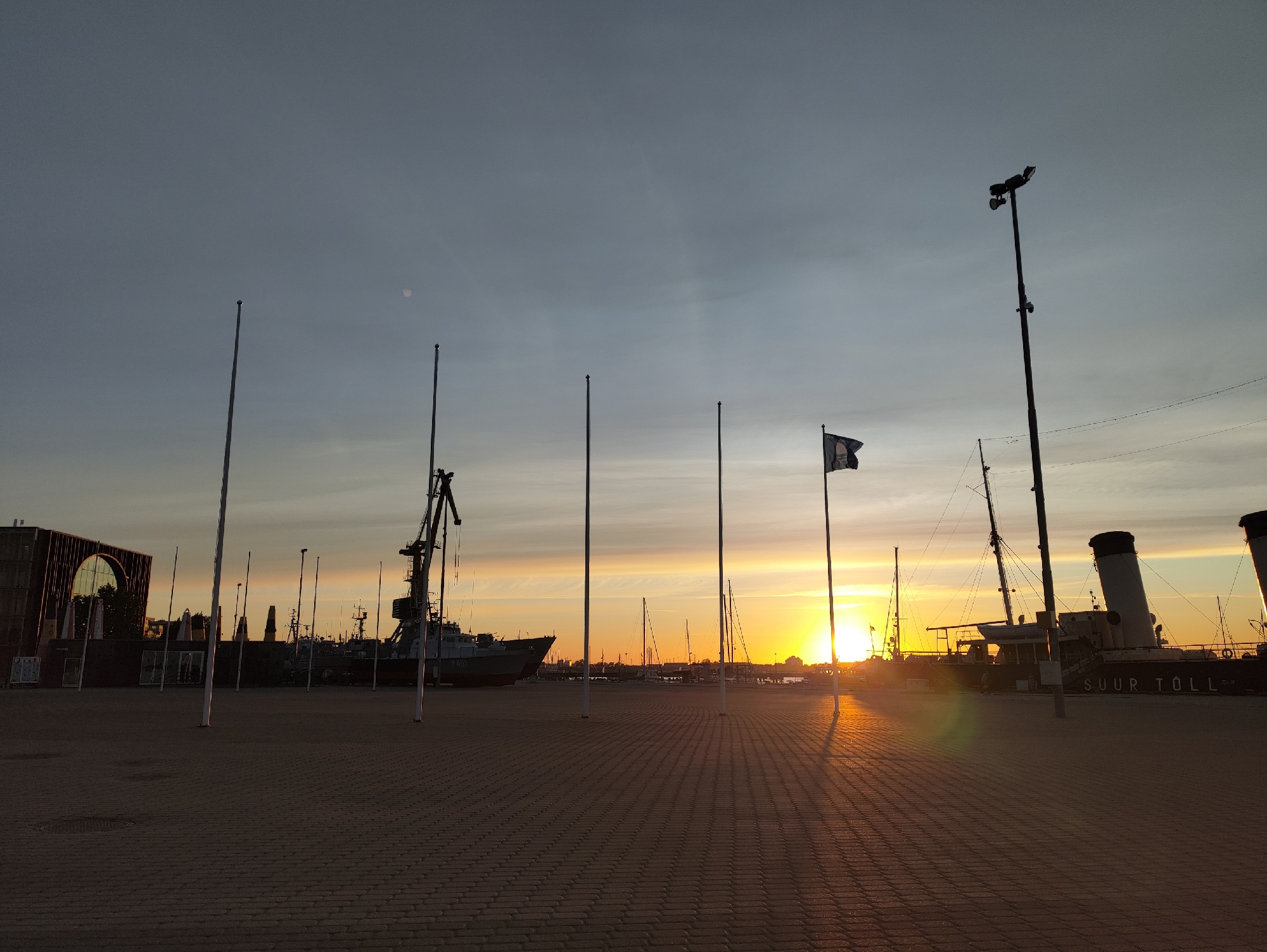 The image captures a sunset scene at a harbor in Tallinn, Estonia. The foreground is paved with brickwork, casting long shadows from several tall, thin poles. In the background, several ships are docked, including a large vessel with the name "SUUR TOLL" visible on its side. A crane is also present, silhouetted against the bright sky. A flag is flying from a flagpole on the right side of the image. The sky is filled with hues of orange, yellow, and blue, indicating the sun is setting. The overall atmosphere is calm and serene.