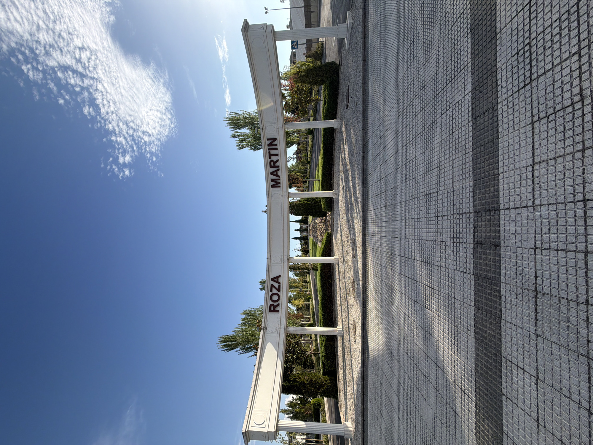 The image shows a wide, arched entrance structure with the words "ROZA MARTIN" inscribed in maroon lettering. The structure is white and features fluted columns. Beyond the arch, a well-maintained park-like area is visible with trees, bushes, and a paved pathway. The ground in the foreground is covered in gray square tiles, with sunlight casting shadows. The sky is blue with scattered white clouds. The location is the Roza Martin Park in Majadahonda, Spain. There are no people or animals visible in the image.