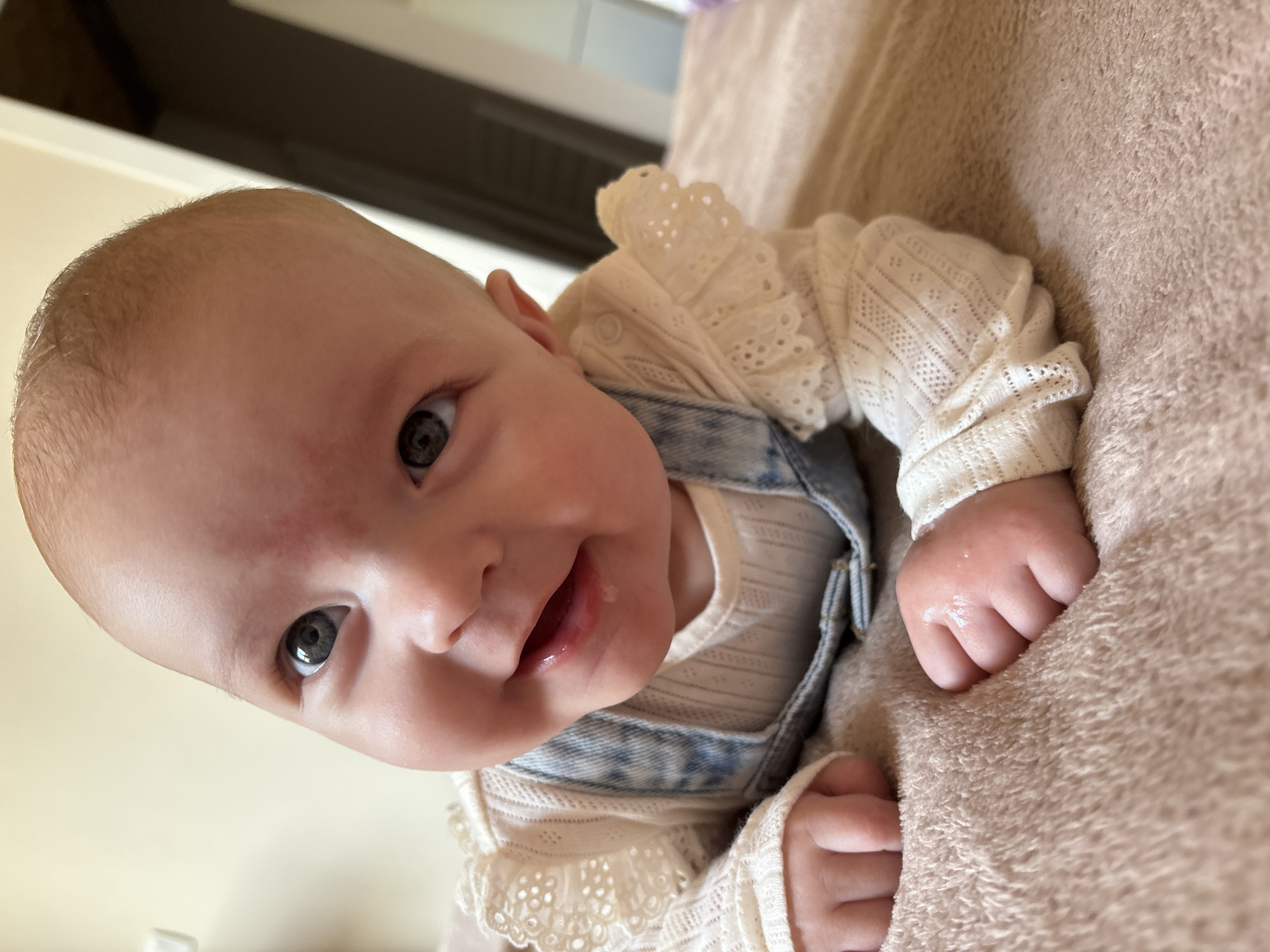 A baby lies on a soft, beige, textured surface. The baby is wearing a white, long-sleeved shirt with a lace collar, underneath light-wash denim overalls. The baby is positioned with its head angled towards the upper left and its body extending towards the lower right of the frame. The baby's eyes are open and looking towards the camera with a slight smile. Saliva is visible at the corner of the baby's mouth. The baby's hands are visible, grasping at the beige surface. Part of a white surface is visible in the upper left. The location is not identifiable as Chaikovsky, Russia.