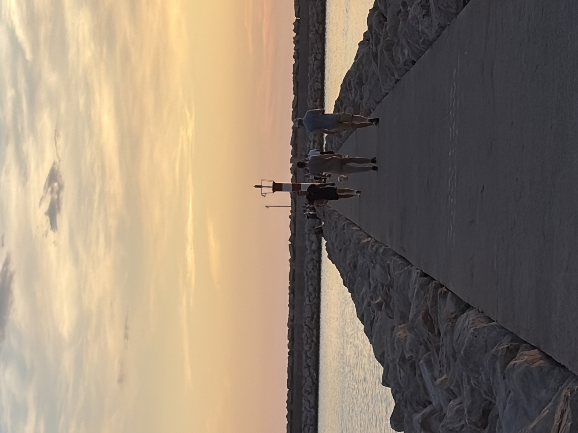 A long concrete pier extends into the water, bordered by large rocks. The pier leads the eye towards a lighthouse-like structure with red and white stripes. People are walking on the pier, moving away from the viewer. The sky is a mix of yellow and orange, indicating sunset. The scene is in Bar, Montenegro.