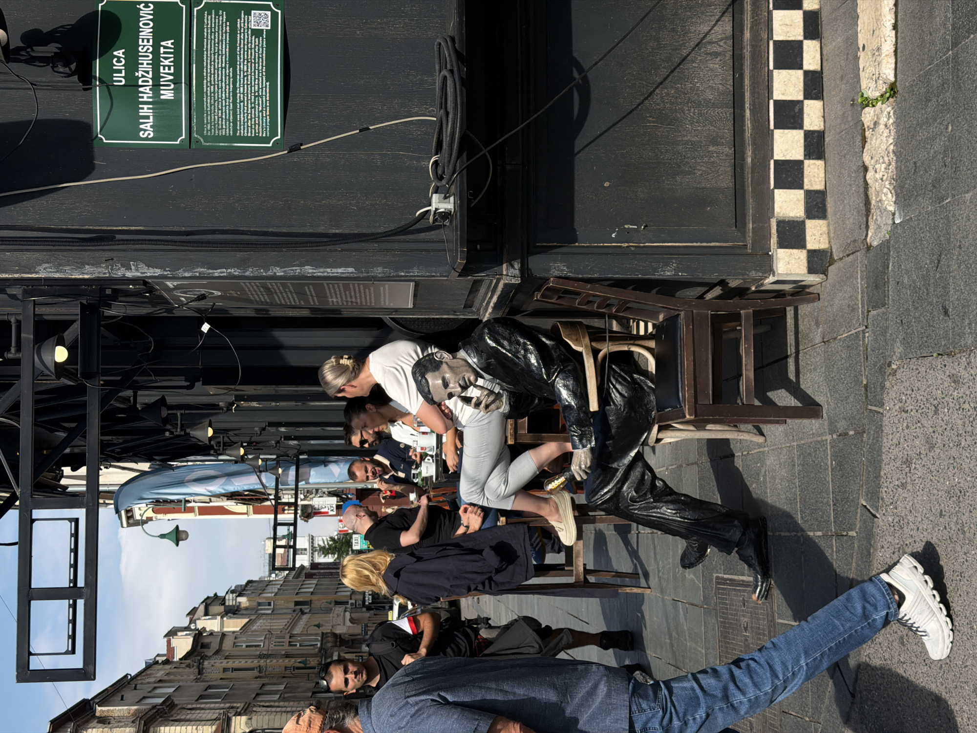 The image depicts a street scene in Sarajevo, Bosnia and Herzegovina, specifically on Ulica Salih Hadžihuseinović Muvekita. In the foreground, a person in blue jeans and white athletic shoes walks on a cobblestone sidewalk. To the right, a life-sized statue of Nikola Tesla, dressed in a black suit, is seated on a chair. The statue is positioned on the sidewalk next to a dark-colored building with a checkered base.
In the background, several people are seated at tables outside a cafe or restaurant. They appear to be engaged in conversation and enjoying beverages. The street is lined with buildings, some of which have historical architectural features. Overhead, there are signs and structural elements related to the businesses. The weather appears to be sunny, casting shadows on the ground. A green street sign with white lettering identifying "ULICA SALIH HADŽIHUSEINOVIĆ MUVEKITA" is prominently displayed on the building.