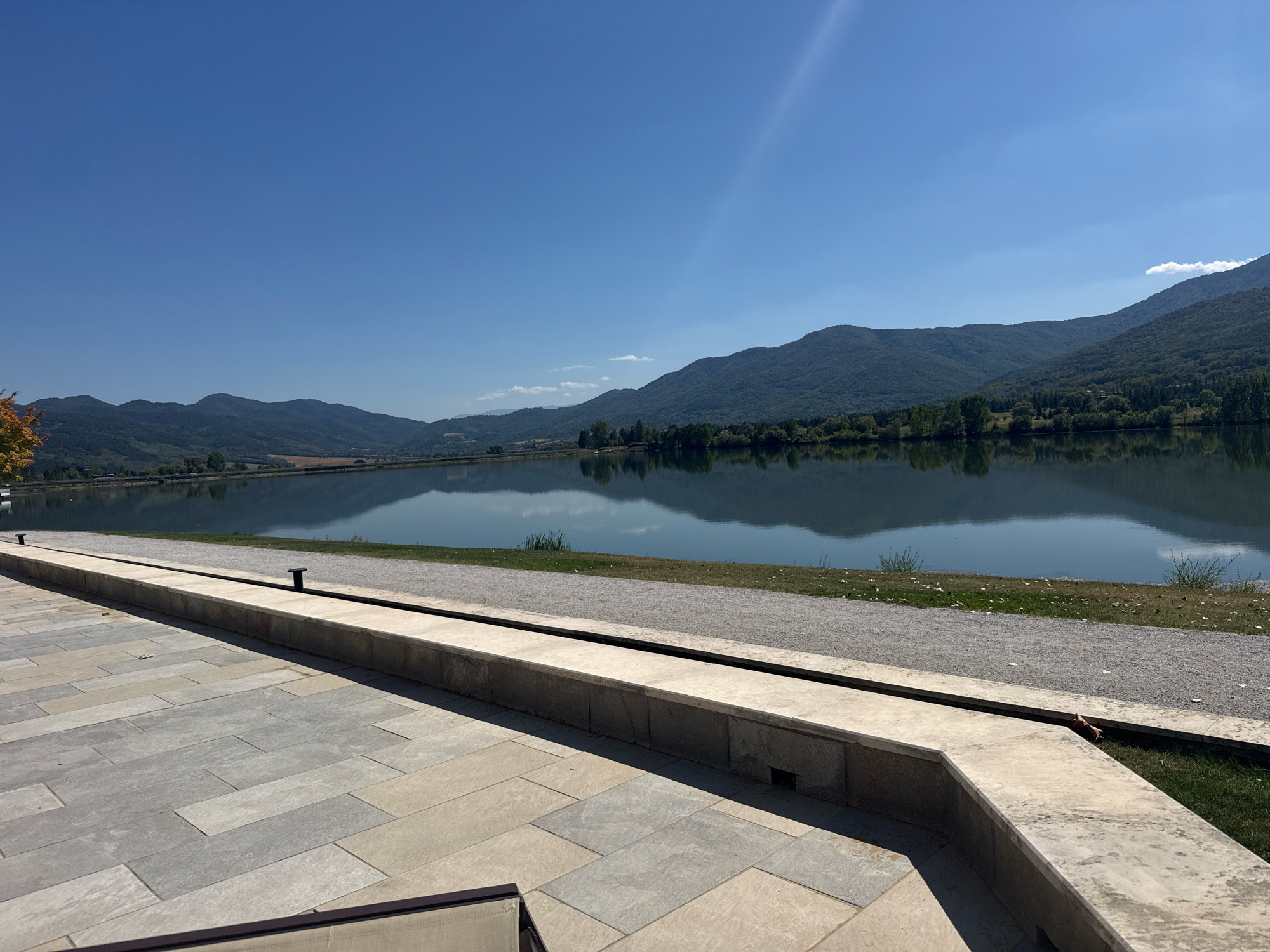 The image depicts a serene waterscape with a large, calm lake reflecting the surrounding mountains under a clear blue sky. The foreground features a paved patio area with large, rectangular stone tiles in varying shades of gray and beige. A low, concrete wall separates the patio from a gravel path and a grassy area bordering the lake.
In the distance, rolling, tree-covered mountains form a picturesque backdrop. The lake itself is smooth, with the reflections of the mountains and trees creating a mirror-like effect. The overall scene suggests a tranquil outdoor setting, possibly a park or a resort area.
The location is identified as the city of Pravets, Bulgaria, known for its lake and surrounding natural beauty. There are no people visible in the image, and the focus is on the natural landscape and the architectural elements of the patio. The lighting suggests a sunny day.