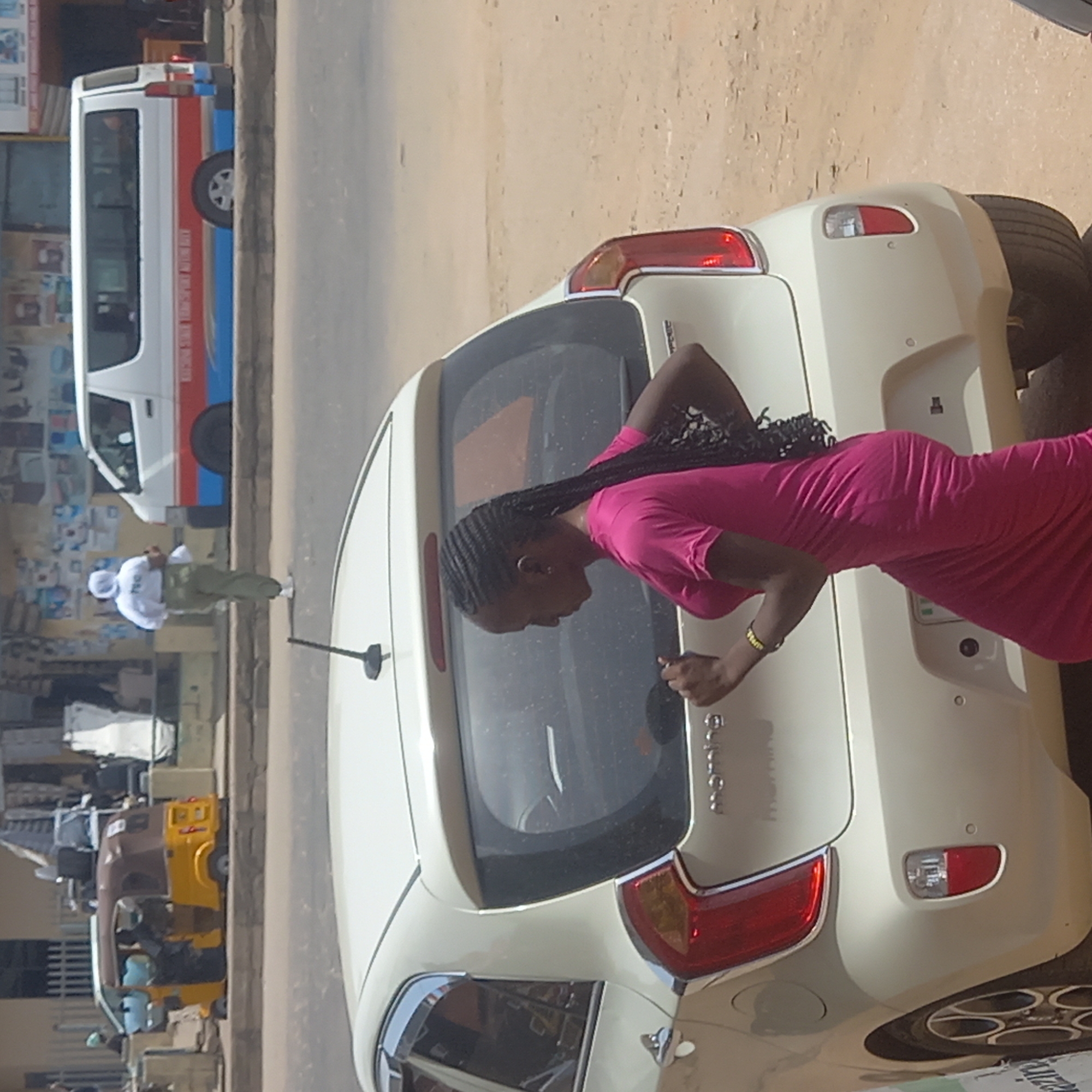 The image shows a woman in a pink dress standing at the rear of a white Kia Morning car on a street in Dadi Riba, Nigeria. The woman is leaning toward the car's trunk, appearing to open or close it. Other vehicles, including a passenger bus with a white, red, and blue paint scheme, and a yellow and brown motorized tricycle (keke napep), are visible in the background. A man in light-colored clothing stands beside a building with displayed goods, suggesting a business or commercial area. The background appears to be a typical street scene with buildings and other objects, indicative of a busy street.