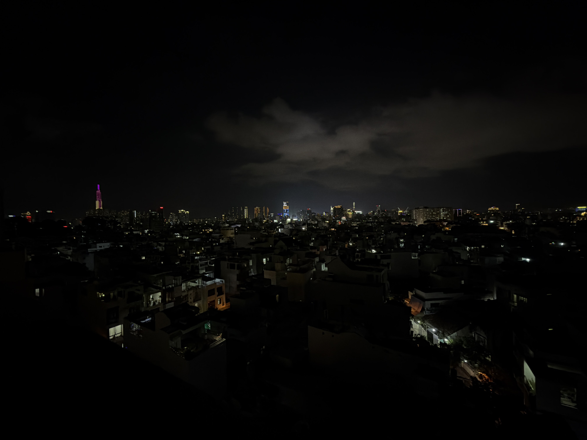 This nighttime image presents a panoramic, high-angle view of a sprawling cityscape, likely from Thủ Đức, Vietnam. The upper half of the frame is dominated by a dark sky, featuring scattered clouds faintly illuminated from below by city lights.
The urban landscape fills the lower half of the photograph, stretching from the immediate foreground to the distant horizon. In the far distance, a prominent cluster of skyscrapers defines the central city skyline. One particularly tall, slender skyscraper on the left is strikingly illuminated in vibrant purple and magenta hues, characteristic of Ho Chi Minh City's Landmark 81. Other high-rise buildings in the distance exhibit various artificial lights, contributing to the urban glow.
Closer to the viewer, the mid-ground and foreground reveal a dense network of lower-rise residential buildings, such as houses and smaller apartment blocks. Many of these structures have illuminated windows, and some feature exterior lights that cast warm glows on their facades and pathways. The intricate pattern of lights across the diverse architecture suggests a vibrant and populous urban environment. No specific human activity or identifiable individuals are visible within the frame. The scene conveys the vastness and nocturnal energy of a major city. There is no visible text in the image.