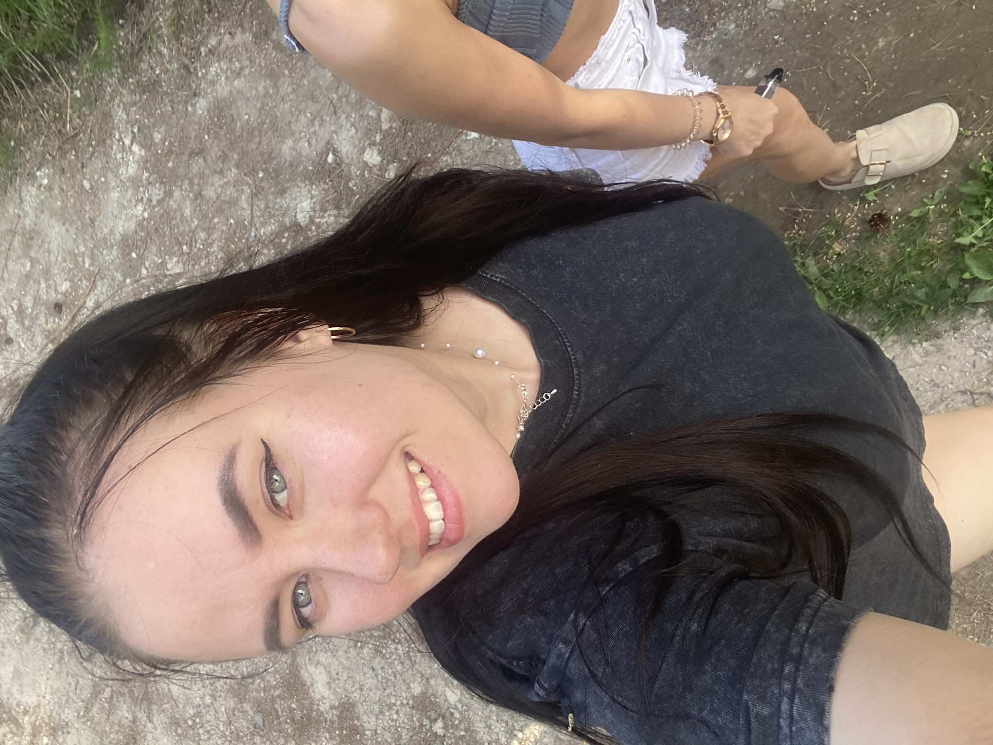 A selfie shows a fair-skinned woman with dark hair and light eyes smiling at the camera. She is wearing a dark-colored t-shirt and a delicate silver necklace. Behind her, another person is visible, wearing a light grey top and white shorts, sitting and holding a small, dark object. They are wearing a gold watch and beige clogs. The ground appears to be a dirt or gravel path with some sparse vegetation. The image was taken outside in natural light. The location of the photograph is the city of Chaikovsky, Russia.