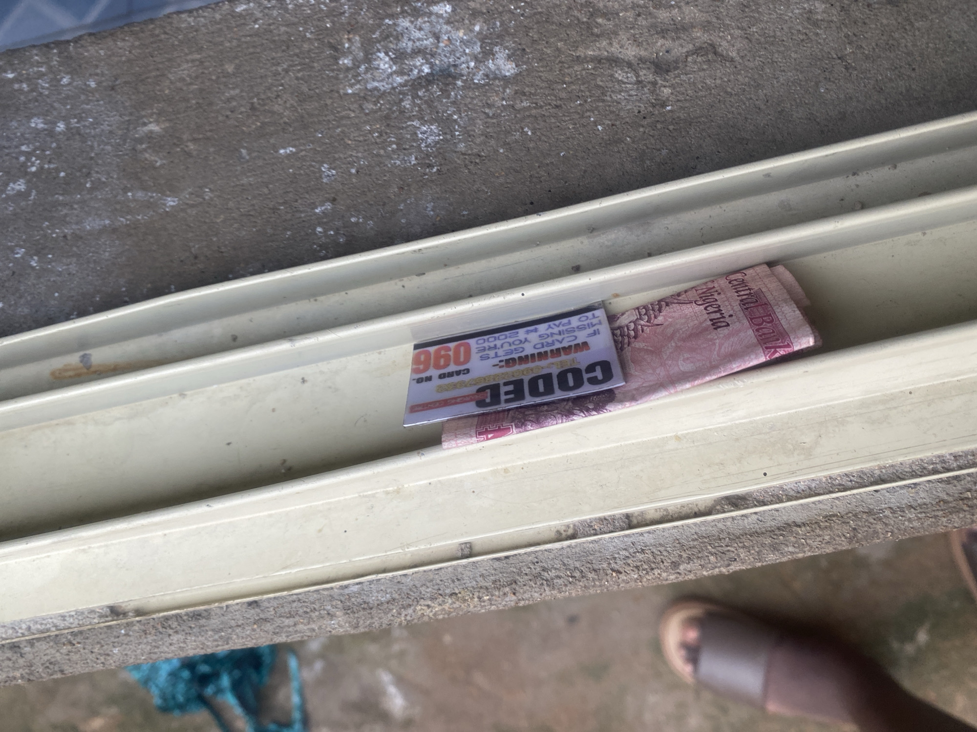 In Jauro Yinu, Nigeria, on a weathered, beige, window frame, a folded pink Nigerian banknote is visible, partially obscured by a white and purple card. The card, featuring a barcode and the word "CODEC" in bold letters, also displays a warning: "IF CARD GETS MISSING YOU'RE TO PAY N 2000." The banknote appears to be from the Central Bank of Nigeria and has text indicating "Nigeria." To the left of the window frame, a person's foot wearing a brown sandal is visible on a concrete surface. A fragment of teal-colored material is also present in the background.