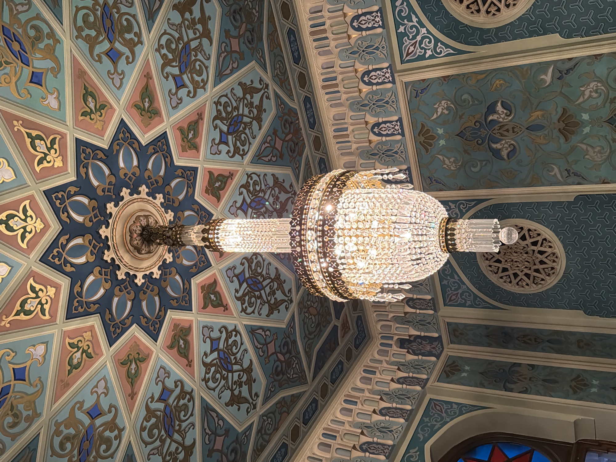 The image captures an upward view of the ceiling within the Orbeliani Presidential Palace located in Tbilisi, Georgia. A prominent feature is a large, ornate chandelier suspended from the center, reflecting light with its numerous crystals. The ceiling displays intricate geometric and floral patterns in shades of blue, gold, and peach, creating a mosaic-like effect. The design incorporates both star-shaped motifs and stylized plant-like elements. The architectural details include a repeating pattern along the edges of the ceiling sections and a circular ventilation grille above the chandelier, contributing to the overall grandeur of the palace's interior.