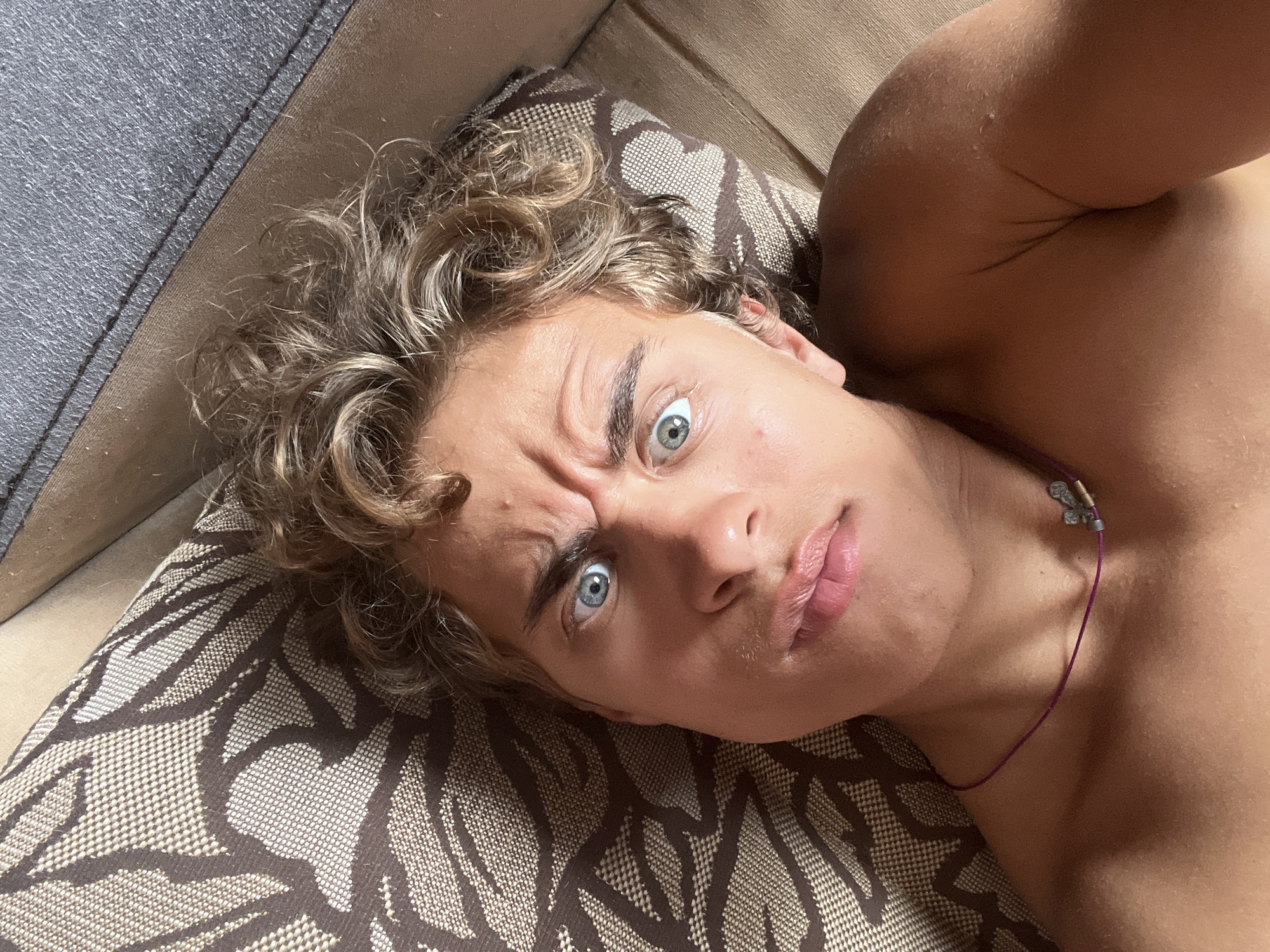A young person with blonde, curly hair and light blue eyes is lying on a patterned couch, looking directly into the camera with a surprised or questioning expression. Their eyebrows are raised, and their mouth is slightly open. The person is shirtless, and a thin purple cord necklace with a small charm is visible around their neck. The background shows the couch cushions and a sliver of a light-colored surface, possibly another furniture item. The lighting suggests an indoor setting, likely during the day. The specific location in Ekibastuz, Kazakhstan cannot be determined from the visual information provided.