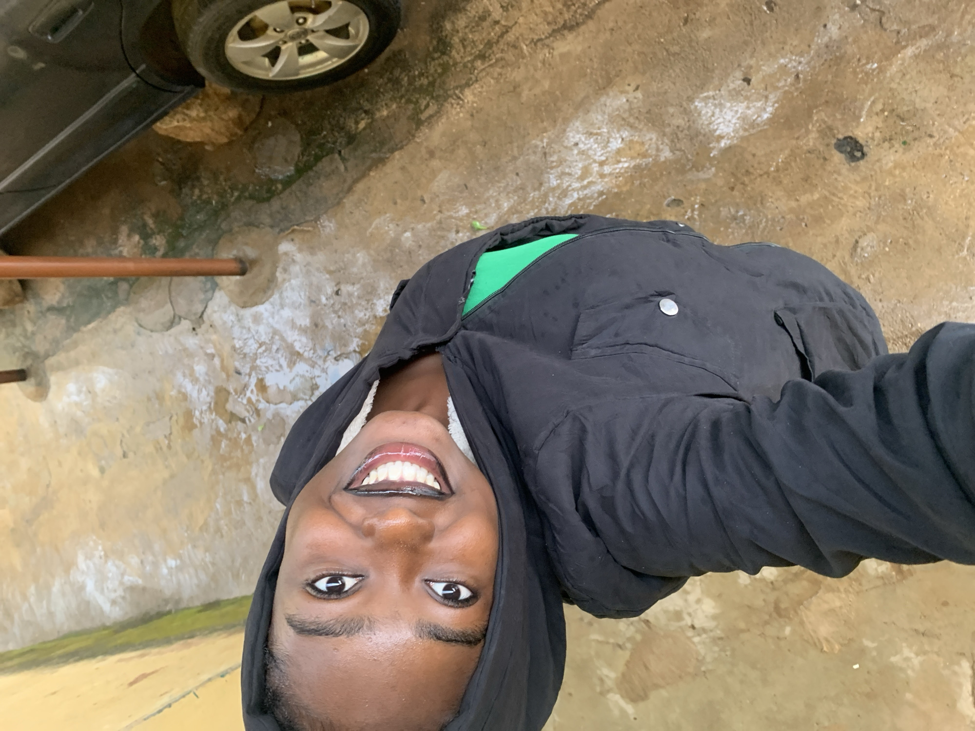 A young woman with dark skin and dark hair, wearing a black hooded jacket over a bright green shirt, smiles directly at the camera. She is outdoors, with a blurry background that includes a portion of a dark-colored car, a concrete ground that appears wet and dirty, and a yellow wall on the left side. A thin, reddish-brown pole stands vertically in the middle ground. The image appears to be taken from a high angle, looking down at the woman. The setting is likely an urban or semi-urban environment.