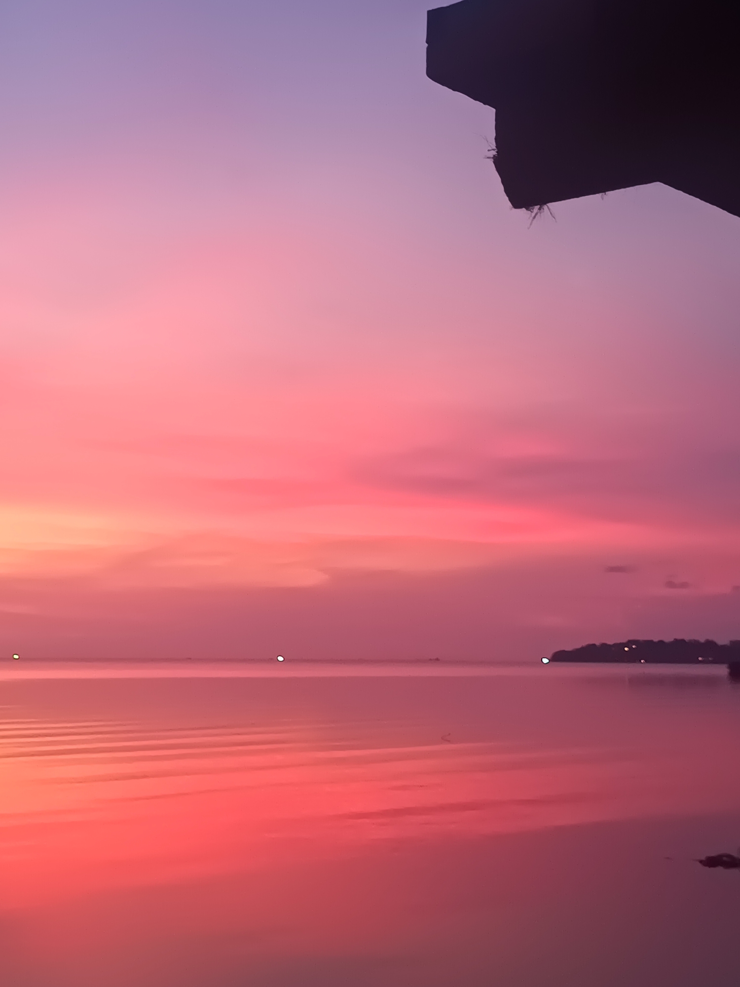 The image depicts a sunset over water, likely a bay or inlet, in Baan Wok Tum, Thailand. The sky transitions from a vibrant pink near the horizon to a lavender hue at the top of the frame. The water reflects these colors, creating a mirrored effect with subtle ripples on the surface. A dark, silhouetted structure is visible in the upper right corner, obscuring part of the sky. In the distance, along the horizon line, there is a faint outline of land with some scattered lights, suggesting a small settlement or development. There are also a few small lights scattered across the water. The overall scene conveys a sense of tranquility and peacefulness.