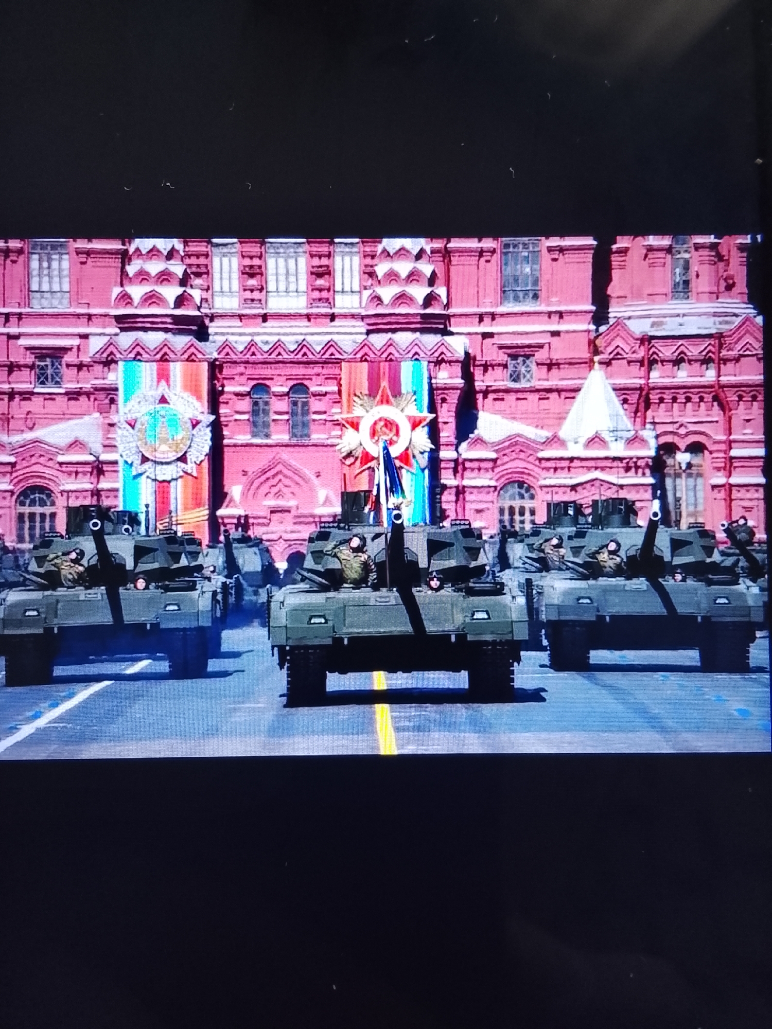The image shows a military parade with several tanks lined up in formation on a street. Behind the tanks is a large, ornate red building with white architectural details and numerous windows. In the center of the parade, there are banners and a large star-shaped medal. The tanks are olive green, and some have soldiers in uniform visible in the open hatches. The street has yellow and white lines painted on it, indicating lanes. The scene appears to be a formal event, likely a national celebration, given the military presence and decorated building. This scene is not in Safonovo, Russia.