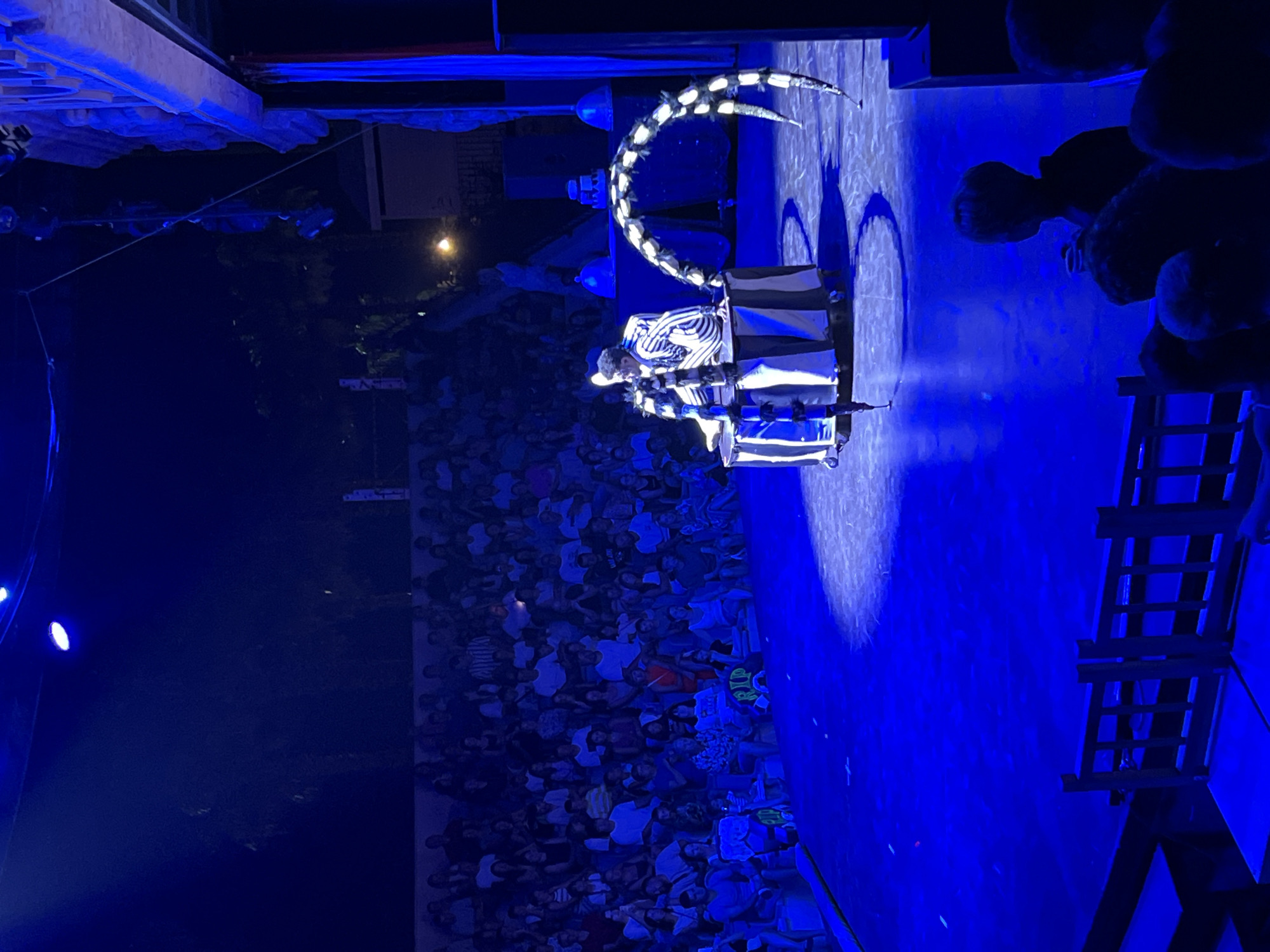 In the city of Serik, Turkey, a stage performance is underway. The stage is illuminated with vibrant blue lights, casting a dramatic glow on the scene. A performer, dressed in a distinctive black and white striped costume, stands on a circular platform. They are interacting with a large, segmented prop that resembles a spider's leg, which arches over them. Behind the performer, an audience is seated in tiered rows, facing the stage. The audience appears engaged, with many individuals looking towards the performance. The overall atmosphere suggests a theatrical or circus-like event.