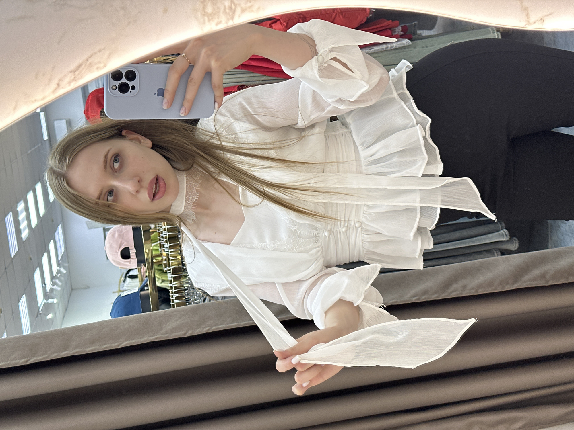 A woman is taking a selfie in a store in Chaikovsky, Russia. She is wearing a white ruffled blouse, black pants, and has long blonde hair. She is holding a light purple iPhone 13 Pro Max in her left hand. The store has clothes racks with various items, and the wall has vertical blinds. The location appears to be a clothing boutique.