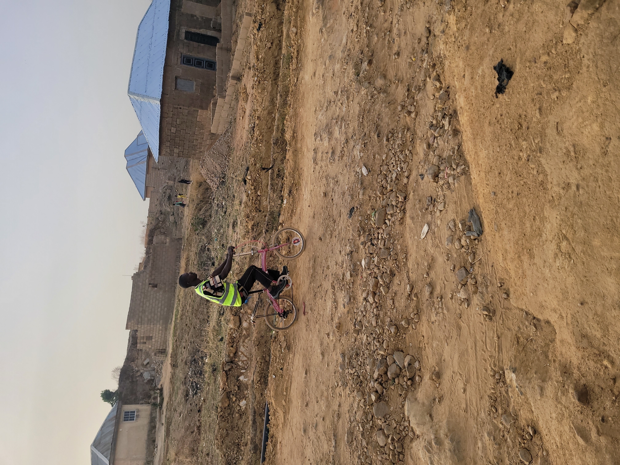 A person wearing a bright yellow vest rides a pink bicycle on a dirt road in Barakin Akawo, Nigeria. The road is lined with rocks and sparse vegetation. Buildings are visible in the background, including unfinished brick structures with blue metal roofs. The sky is overcast.