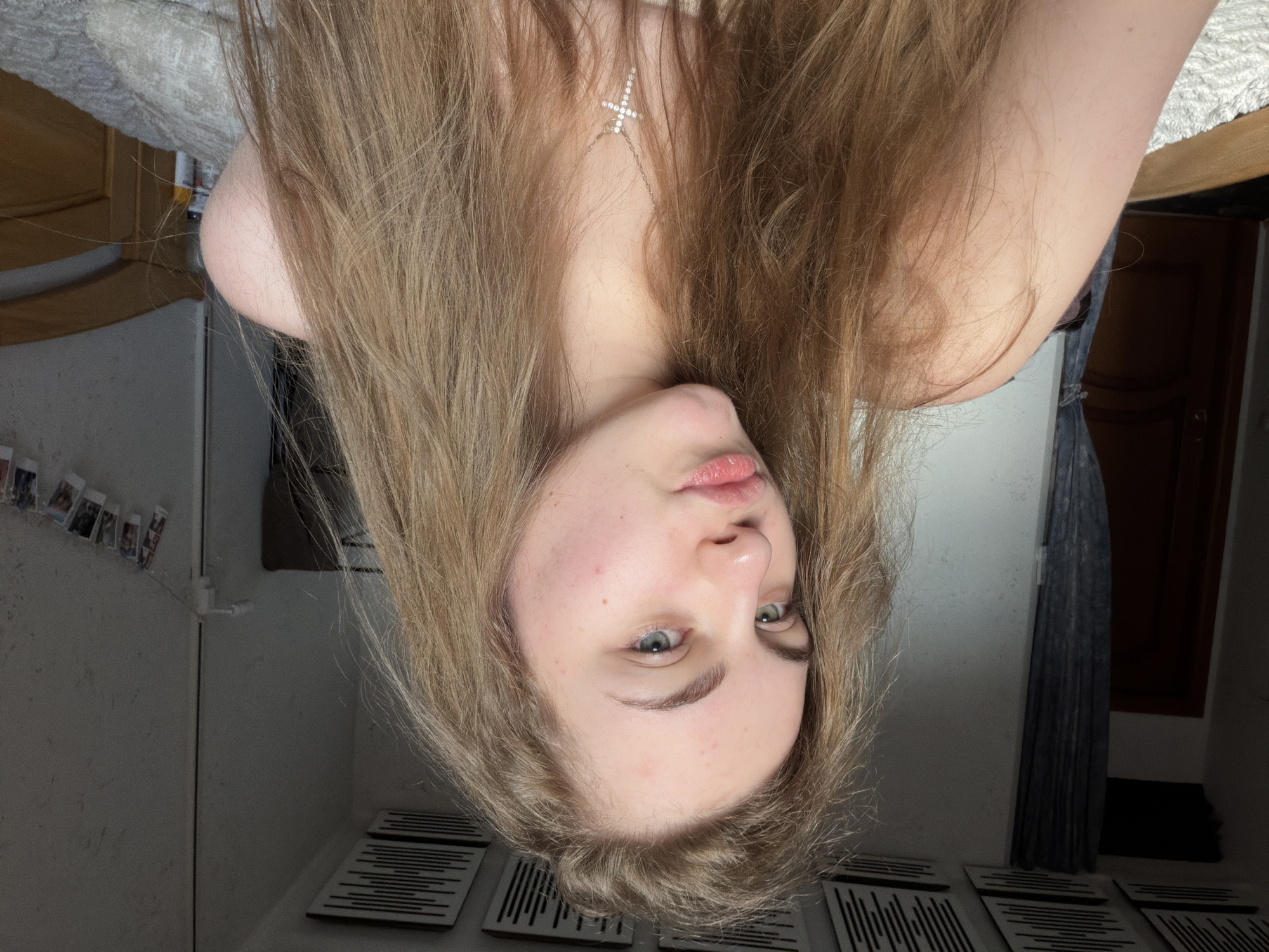 A young woman with long, blonde hair is posing for a selfie in a dimly lit bedroom. She is wearing a necklace with a cross pendant. The room appears to be cluttered, with a bed visible in the background and some framed photos hanging on the wall. The overall lighting suggests it is either early morning or late evening. The location is not specified in the image or associated metadata.