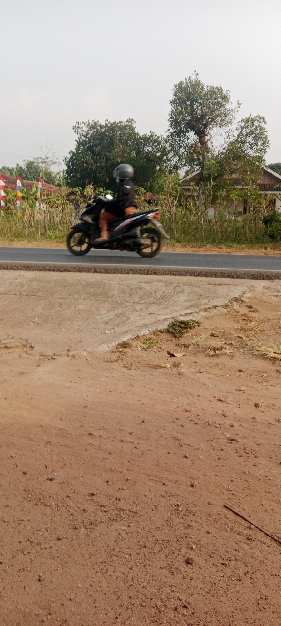 A person wearing a gray helmet and a dark jacket is riding a black scooter on a paved road. The scooter is in motion, indicated by the blur of its wheels. The rider is wearing light brown pants and their feet are visible. The road is bordered by dry grass and dirt on the right side. In the background, there are trees, some with leaves and others bare, and a house with a red roof. There are also flags or banners on the left side of the image, possibly indicating a local event. The sky is overcast and bright. The image was taken from a low angle, looking slightly upwards. The scene appears to be in a rural or semi-rural area, consistent with the location of Gondang Rejo, Indonesia.