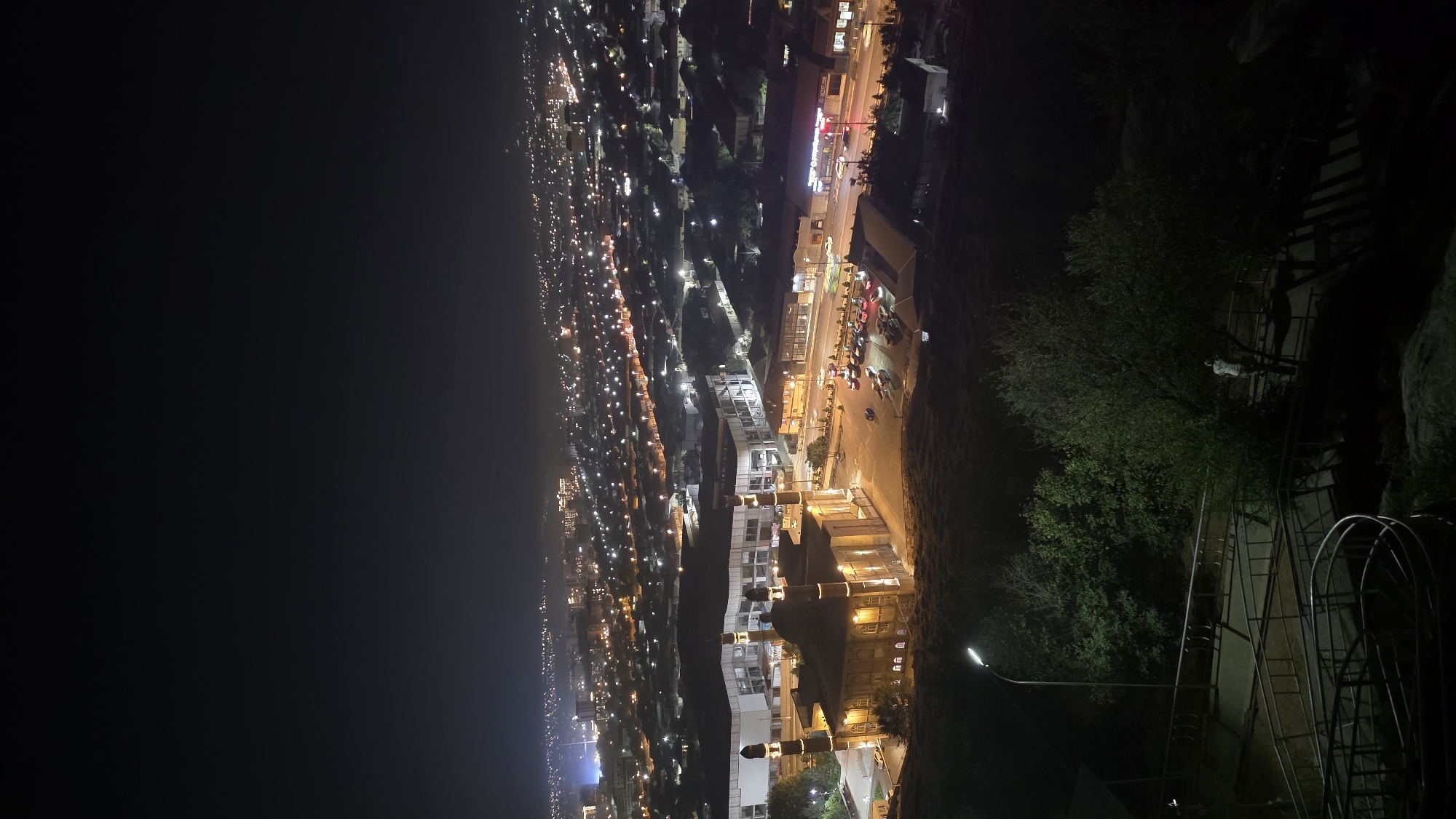 This is a nighttime, high-angle photograph of the city of Osh, Kyrgyzstan, featuring the Sulayman-Too Sacred Mountain National Park in the foreground and the city lights in the background. The focal point in the mid-ground is a well-lit mosque with multiple domes and tall minarets, situated on a grassy slope. Adjacent to the mosque is a large, modern building with illuminated windows. A parking lot filled with cars is visible in front of these structures. The foreground shows a dark, grassy hillside with a paved pathway and metal railings, leading downwards. A single person is visible on the pathway, standing near trees. The background is a sprawling cityscape with numerous lights indicating buildings and streetlights extending into the distance under a dark night sky.